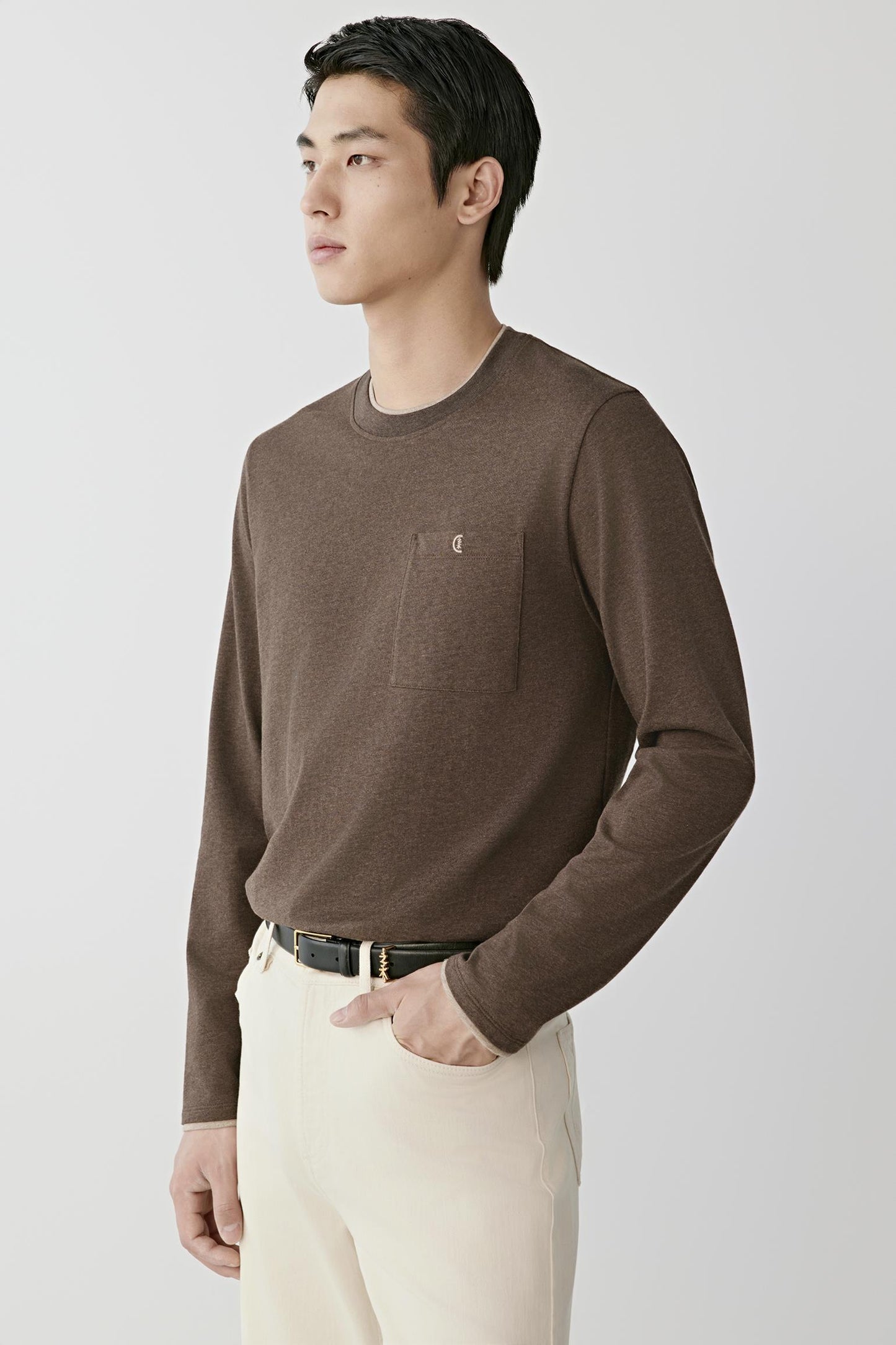 A man wearing a brown long-sleeve crewneck sweater with a chest pocket and cream trousers, standing against a light gray studio background.