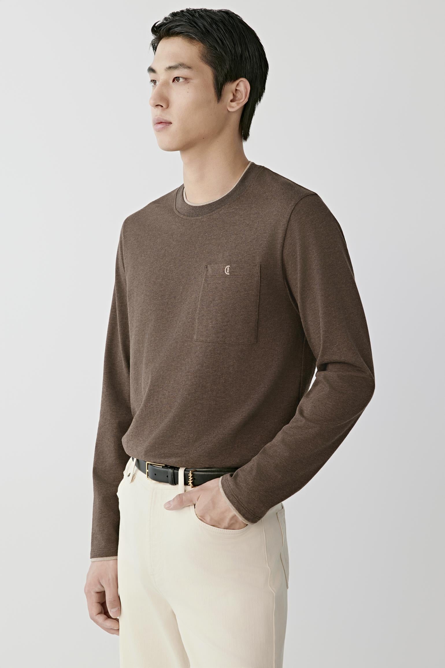 A man wearing a brown long-sleeve crewneck sweater with a chest pocket and cream trousers, standing against a light gray studio background.