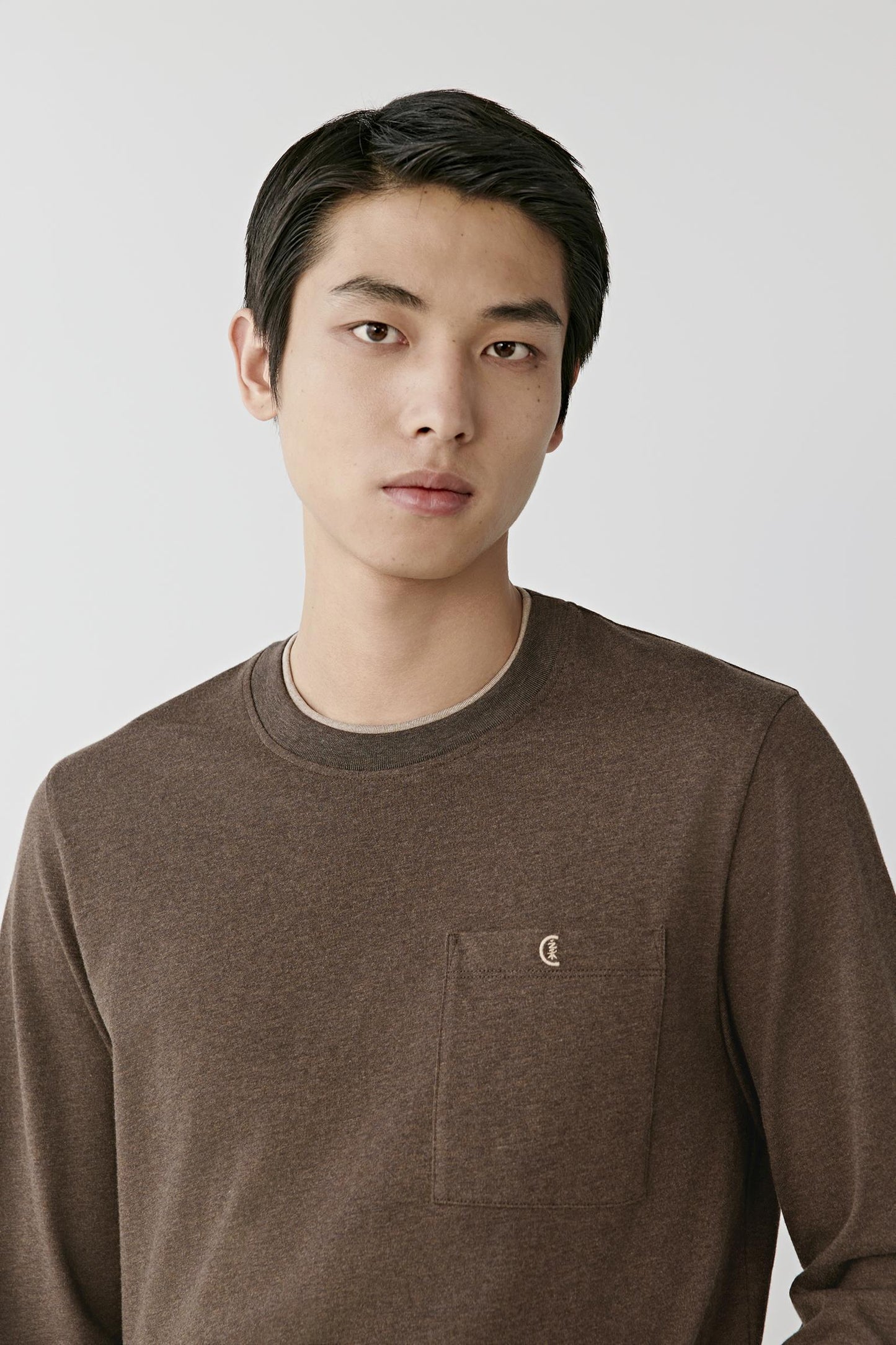 A man wearing a brown crewneck long-sleeve knit sweater with a chest pocket and a small embroidered logo, photographed against a light gray studio background.