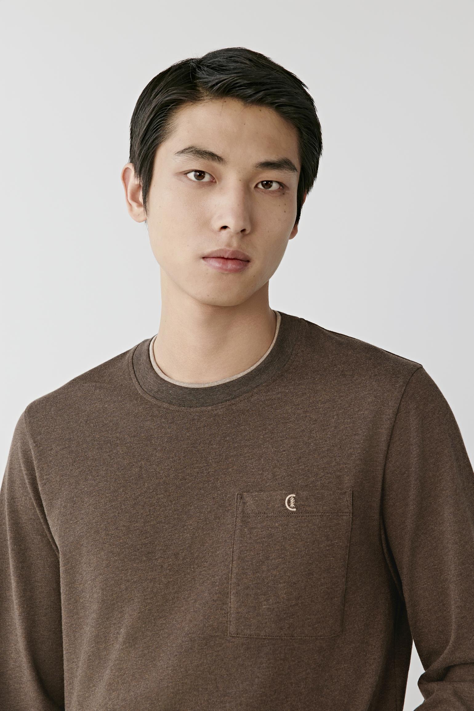 A man wearing a brown crewneck long-sleeve knit sweater with a chest pocket and a small embroidered logo, photographed against a light gray studio background.