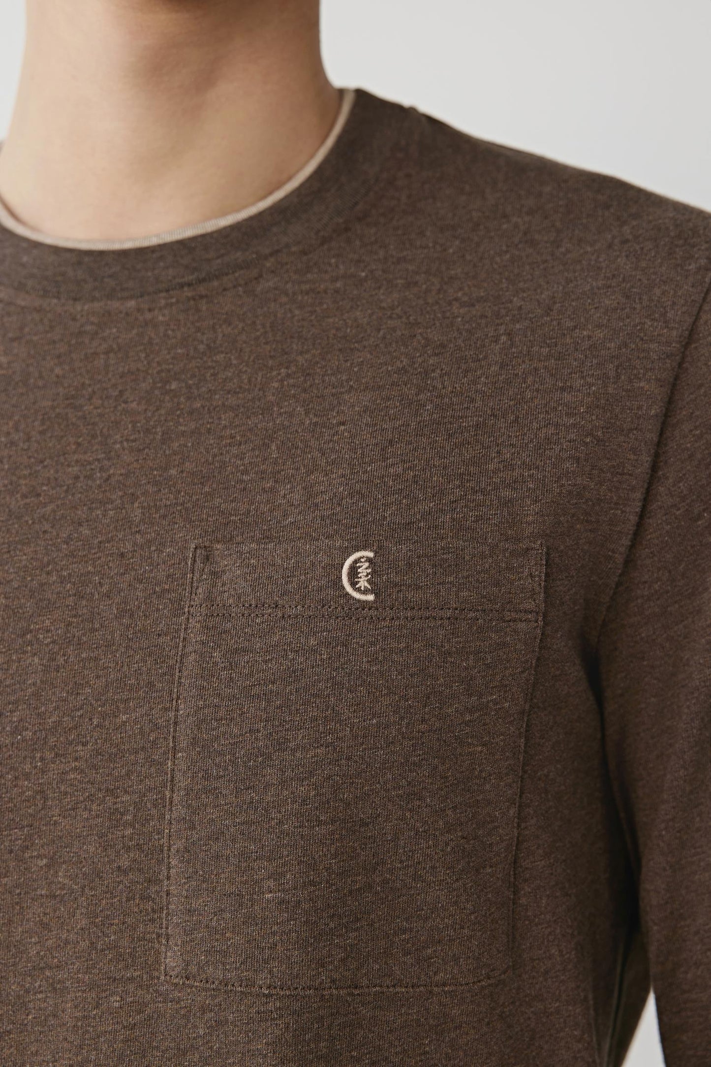 Close-up of a person wearing a dark brown long-sleeve crewneck shirt with a chest pocket and a small embroidered crescent moon logo on the pocket.