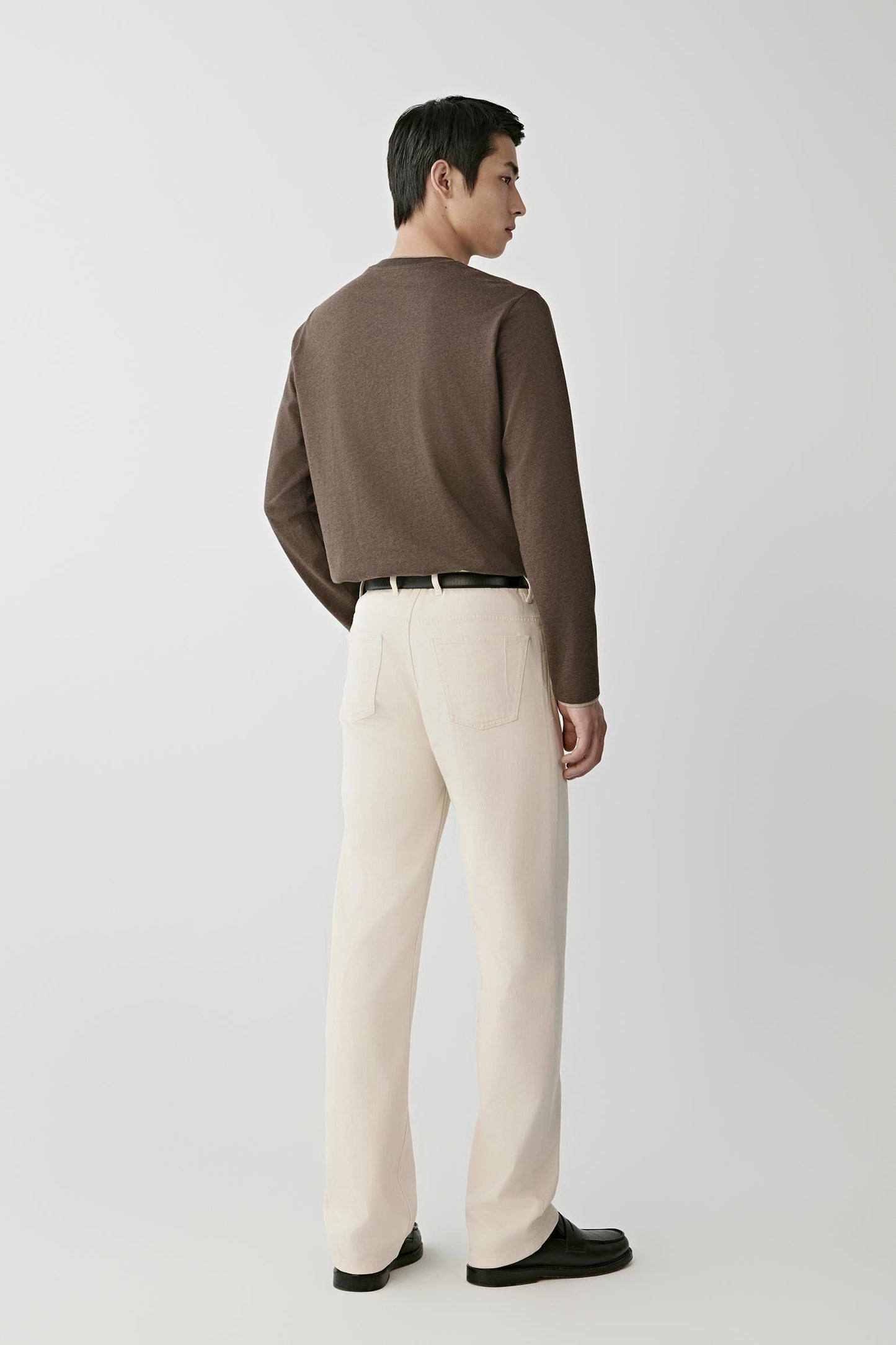 A man shown from the back wearing a brown knit sweater, off-white trousers, a black belt, and black loafers, standing against a light gray studio background.