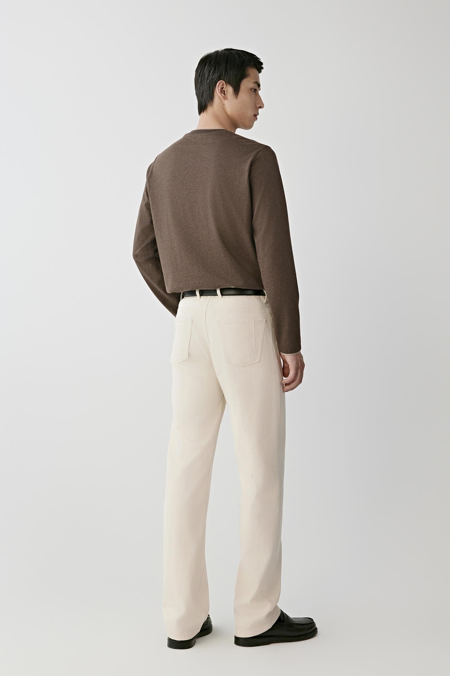 A man shown from the back wearing a brown knit sweater, off-white trousers, a black belt, and black loafers, standing against a light gray studio background.
