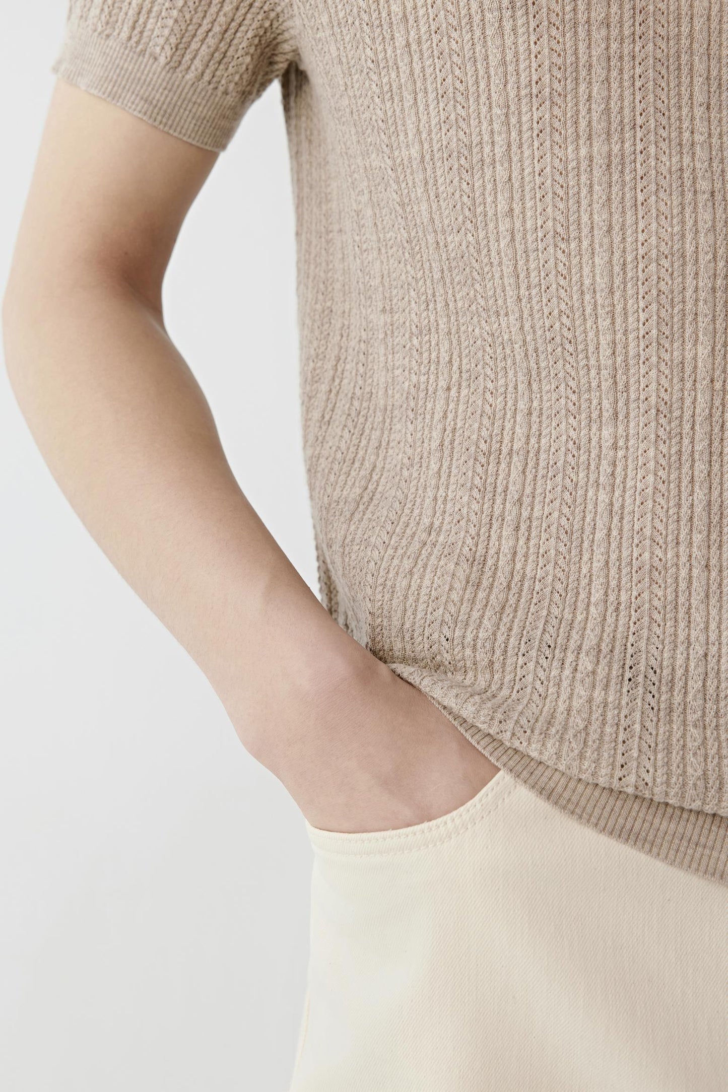 A man modeling a light beige short-sleeve knit polo shirt, paired with cream trousers, a black belt, and off-white sneakers, against a plain light gray studio background.