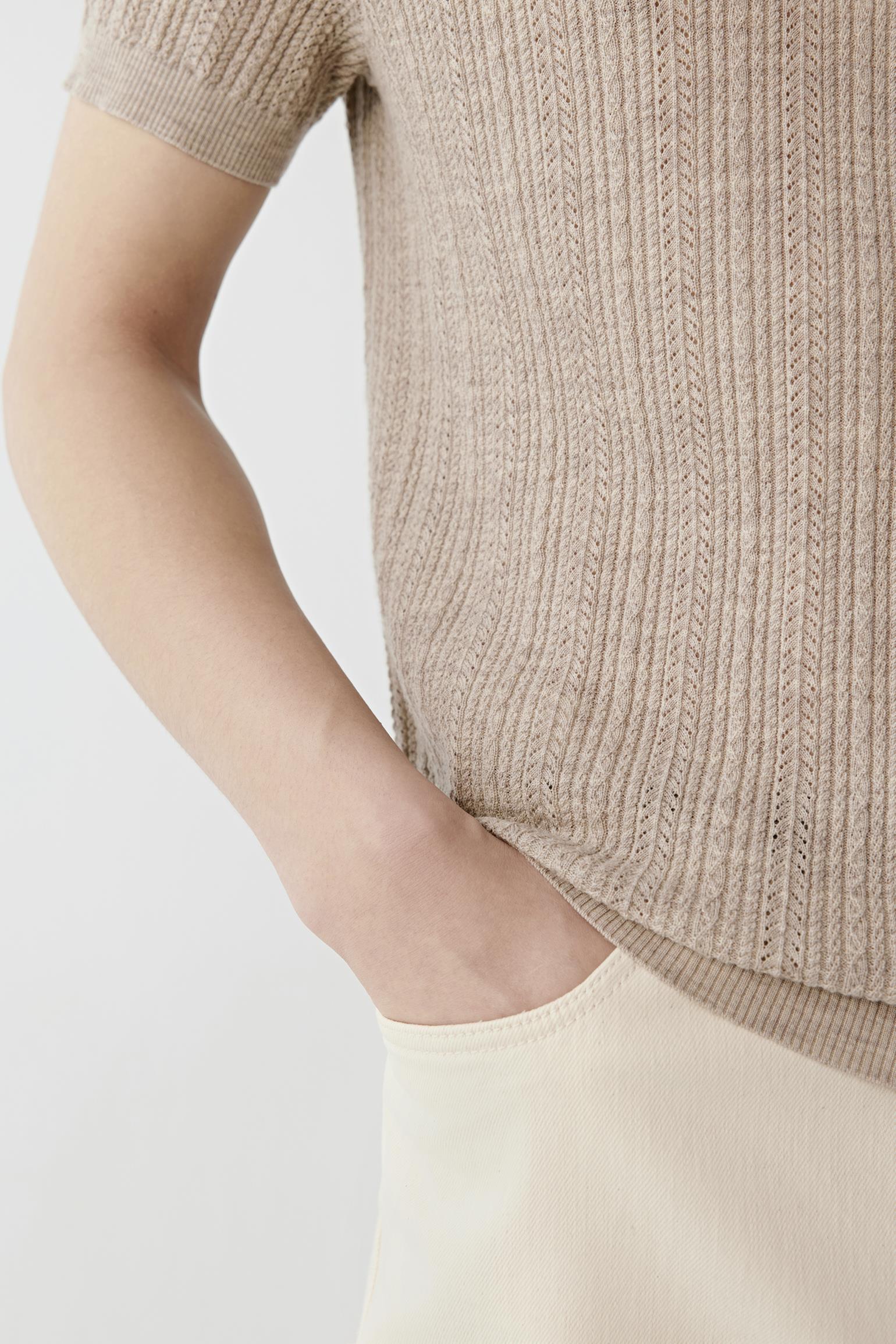 A man modeling a light beige short-sleeve knit polo shirt, paired with cream trousers, a black belt, and off-white sneakers, against a plain light gray studio background.