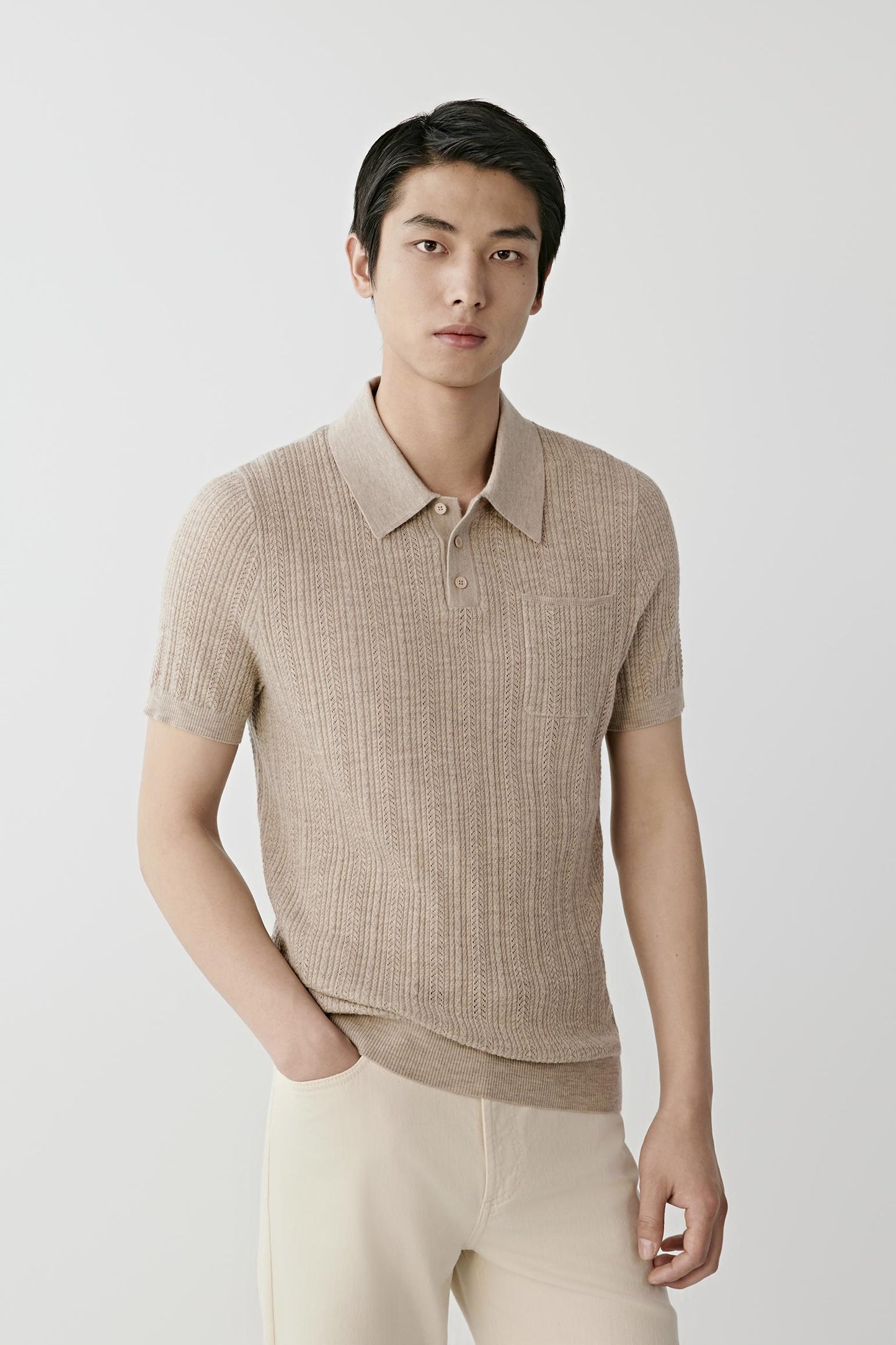 A man modeling a light beige short-sleeve knit polo shirt, paired with cream trousers, a black belt, and off-white sneakers, against a plain light gray studio background.