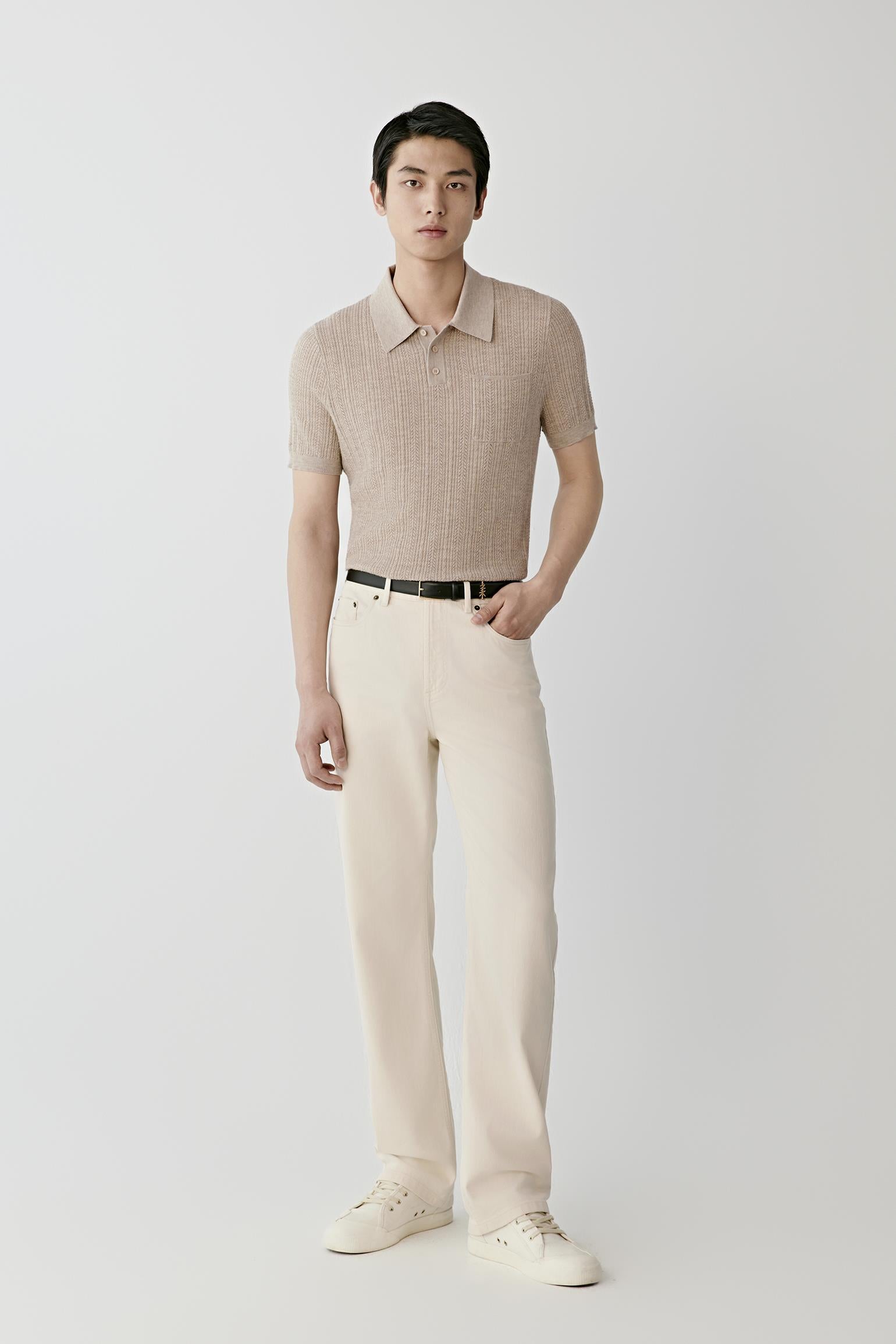 A man modeling a light beige short-sleeve knit polo shirt, paired with cream trousers, a black belt, and off-white sneakers, against a plain light gray studio background.
