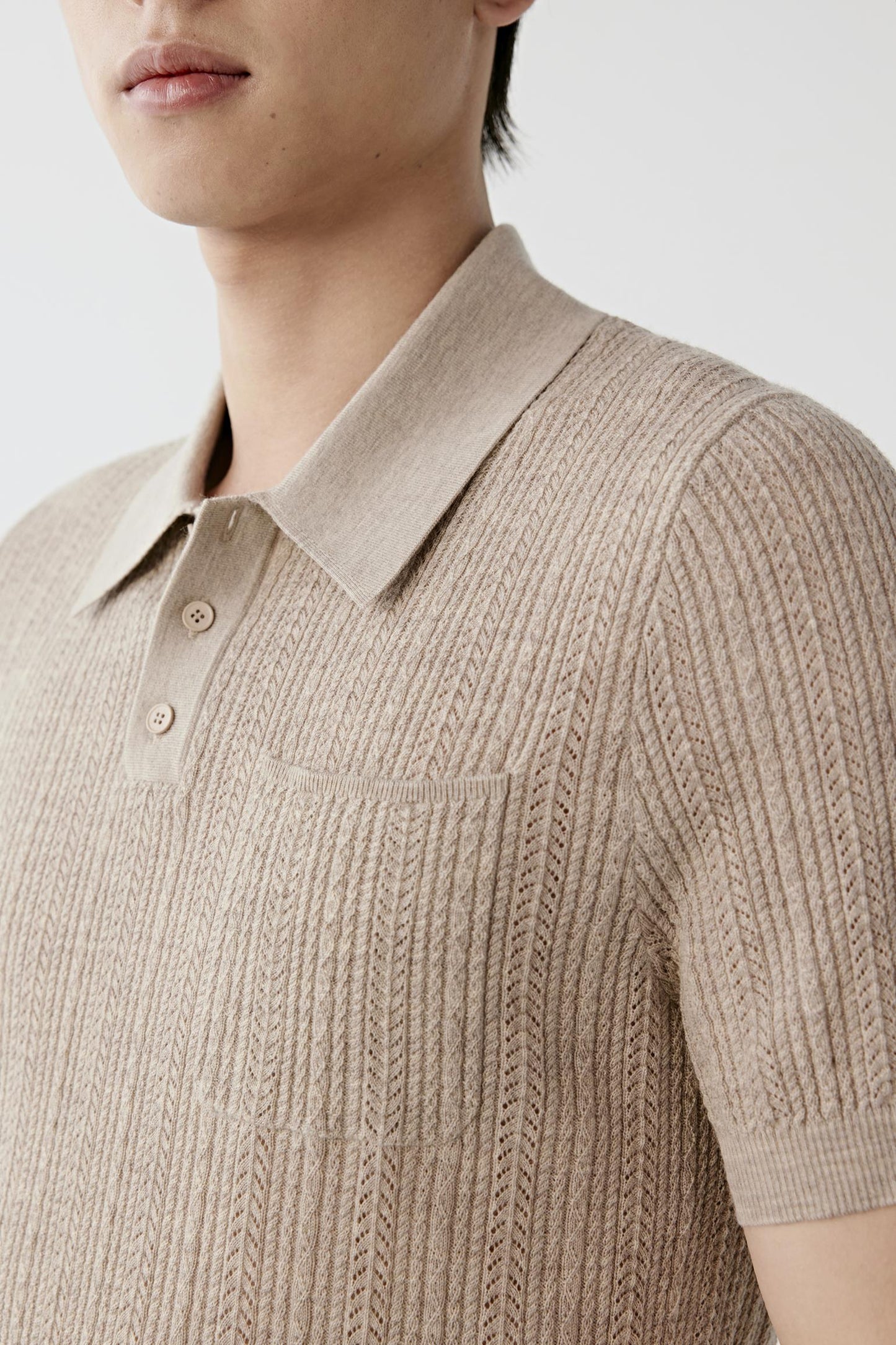 A man modeling a light beige short-sleeve knit polo shirt, paired with cream trousers, a black belt, and off-white sneakers, against a plain light gray studio background.