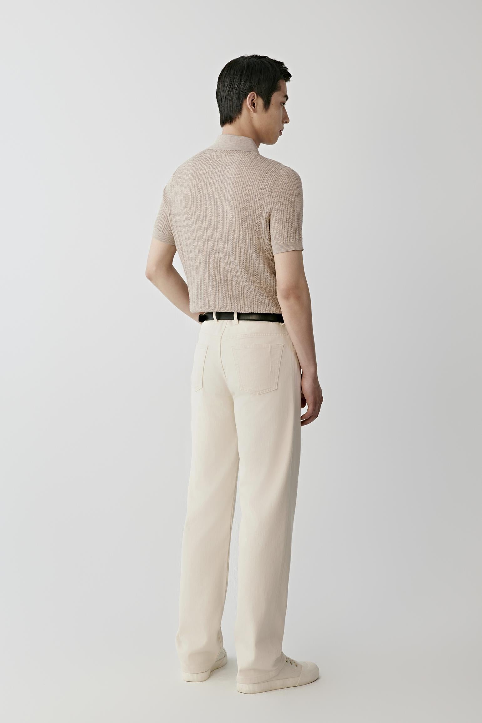 A man modeling a light beige short-sleeve knit polo shirt, paired with cream trousers, a black belt, and off-white sneakers, against a plain light gray studio background.