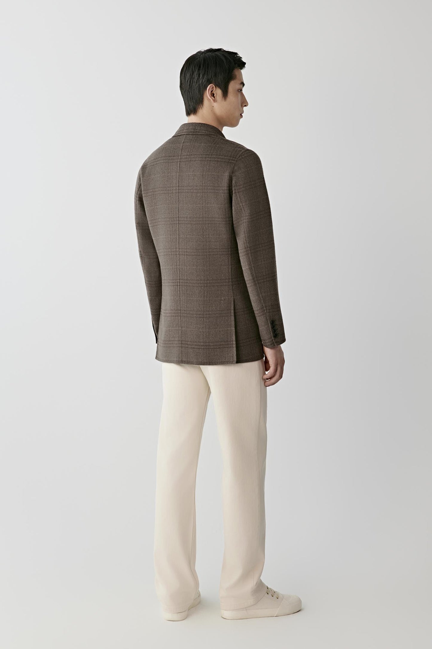 A man shown from the back wearing a brown plaid blazer, cream trousers, and white sneakers, standing against a light gray studio background.