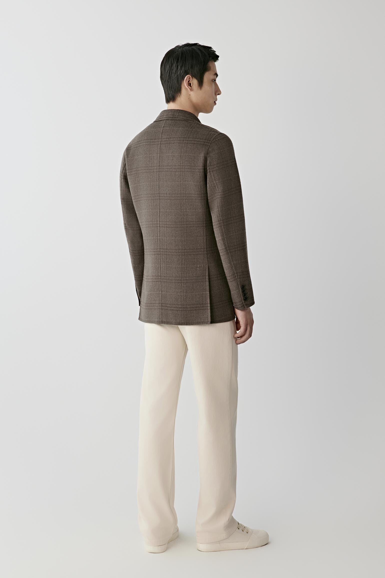 A man shown from the back wearing a brown plaid blazer, cream trousers, and white sneakers, standing against a light gray studio background.