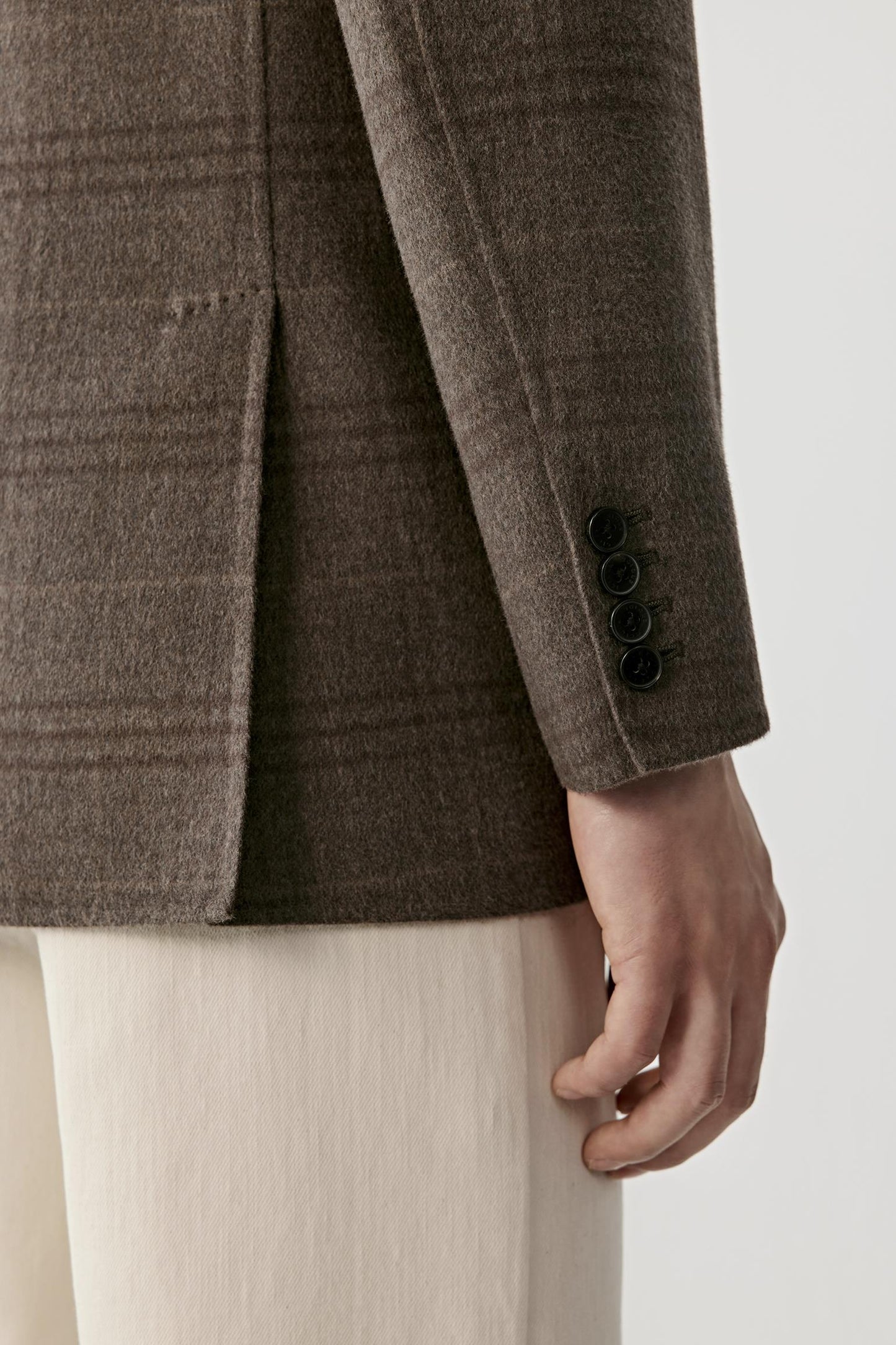 Close-up of a person wearing a brown plaid blazer with black buttons on the cuff, paired with light cream trousers, against a plain light background.