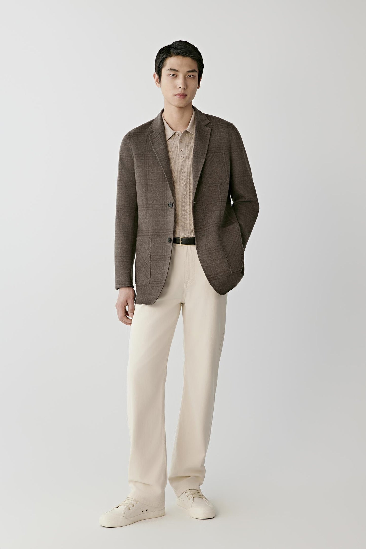A man modeling a brown plaid blazer over a beige collared shirt, paired with cream trousers and white sneakers, against a light gray studio background.