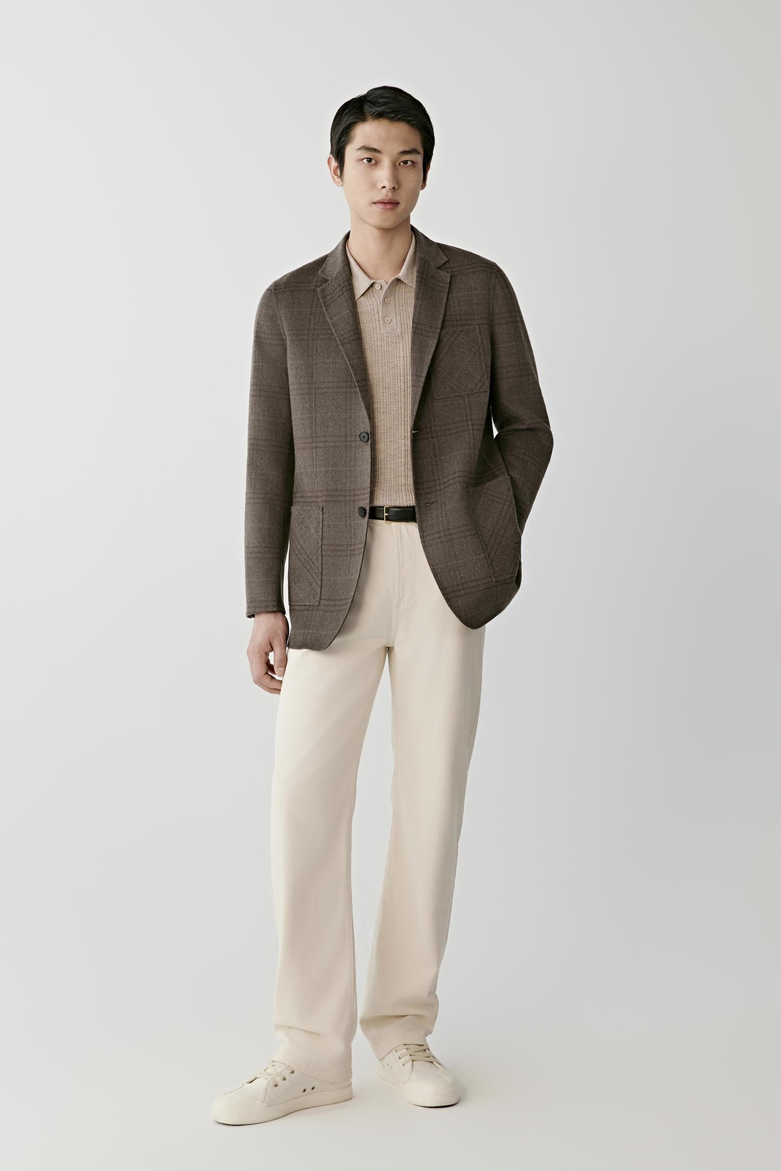 A man modeling a brown plaid blazer over a beige collared shirt, paired with cream trousers and white sneakers, against a light gray studio background.