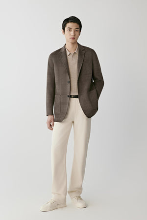 A man modeling a brown plaid blazer over a beige collared shirt, paired with cream trousers and white sneakers, against a light gray studio background.