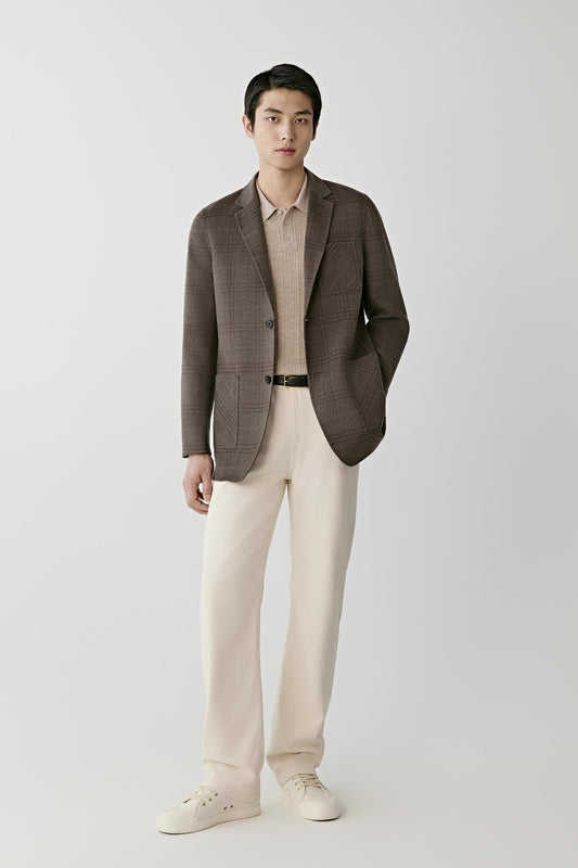 A man modeling a brown plaid blazer over a beige collared shirt, paired with cream trousers and white sneakers, against a light gray studio background.