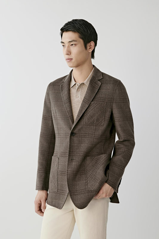 A man wearing a brown plaid blazer over a beige knit polo shirt and cream trousers, standing against a light gray studio background.