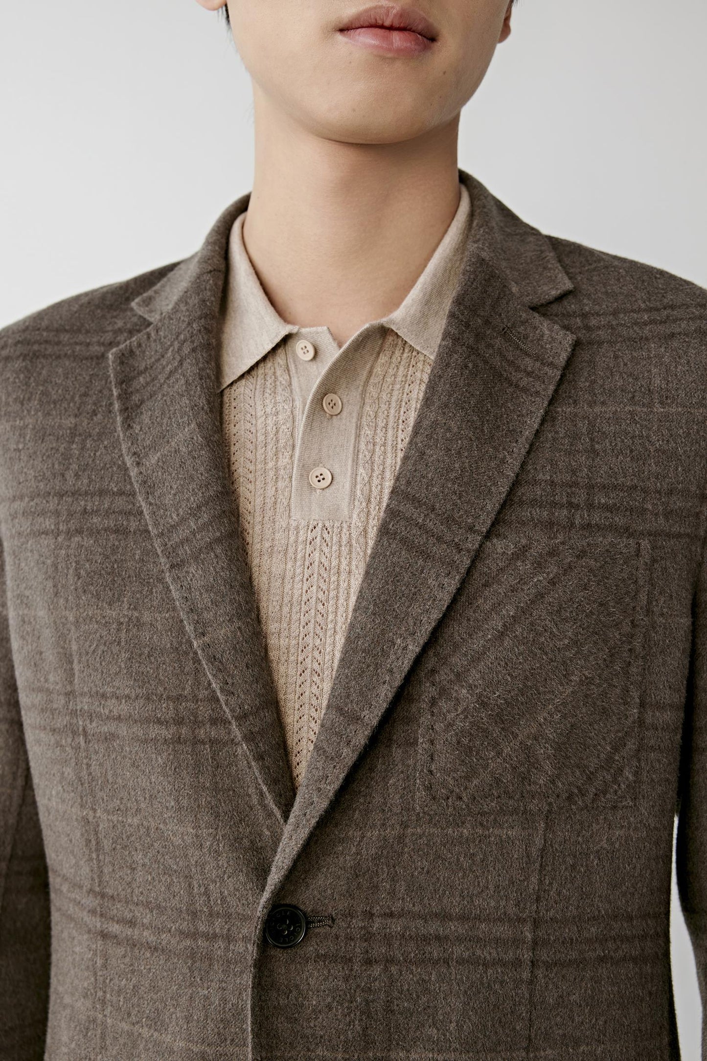 A close-up of a model wearing a brown plaid blazer over a beige knit polo shirt, cropped from the lower face to the waist against a light gray studio background.