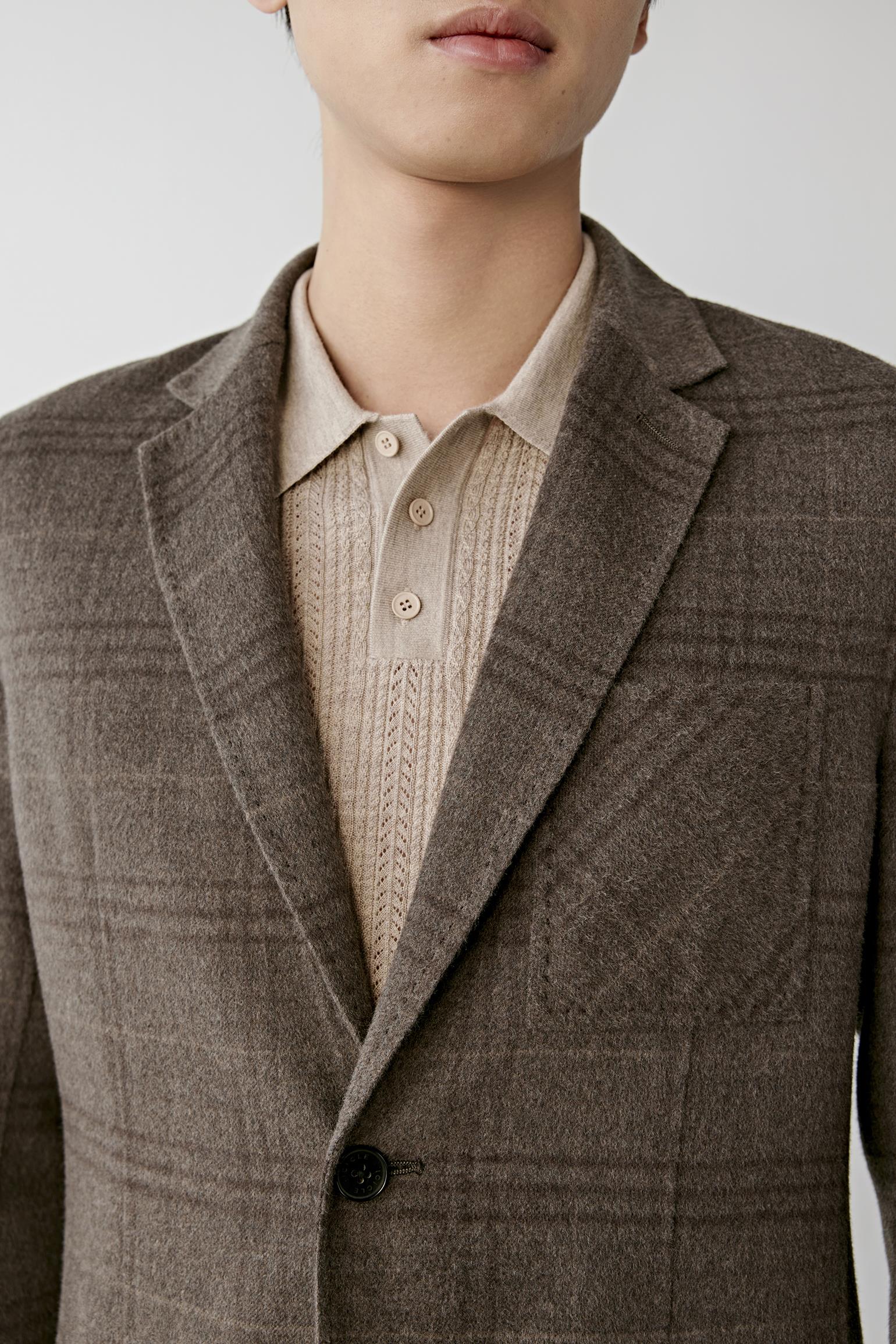 A close-up of a model wearing a brown plaid blazer over a beige knit polo shirt, cropped from the lower face to the waist against a light gray studio background.