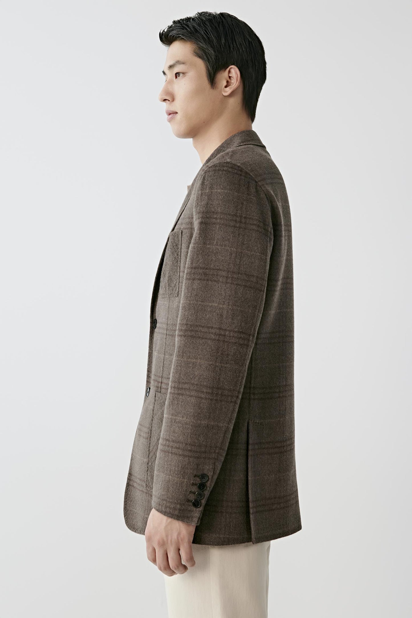 A man shown in profile wearing a brown plaid blazer and cream trousers, photographed against a clean light gray studio background.