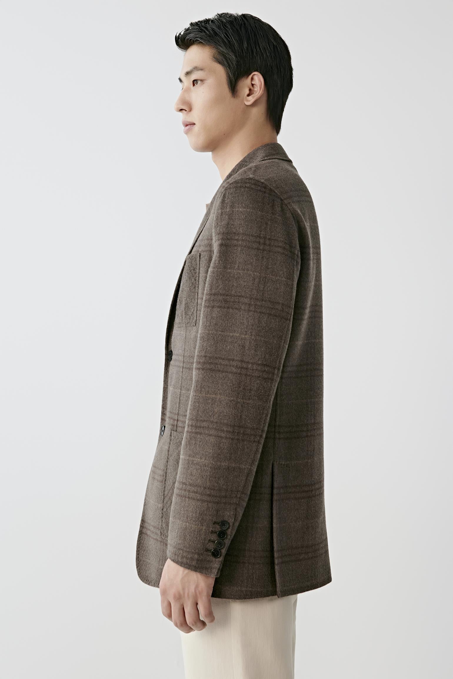 A man shown in profile wearing a brown plaid blazer and cream trousers, photographed against a clean light gray studio background.