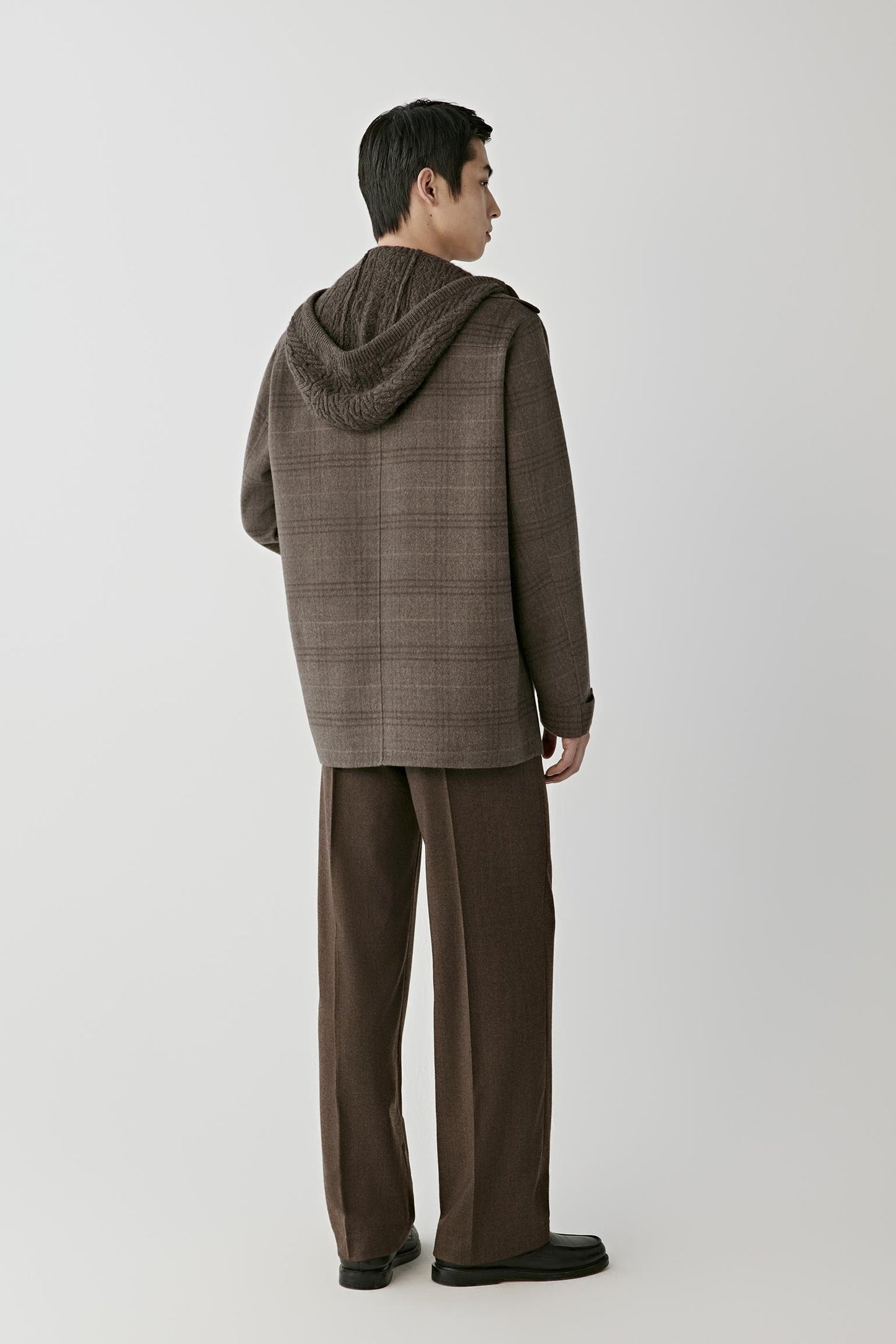 A man shown from the back wearing a brown textured hooded jacket and matching brown trousers, standing against a light gray studio background.