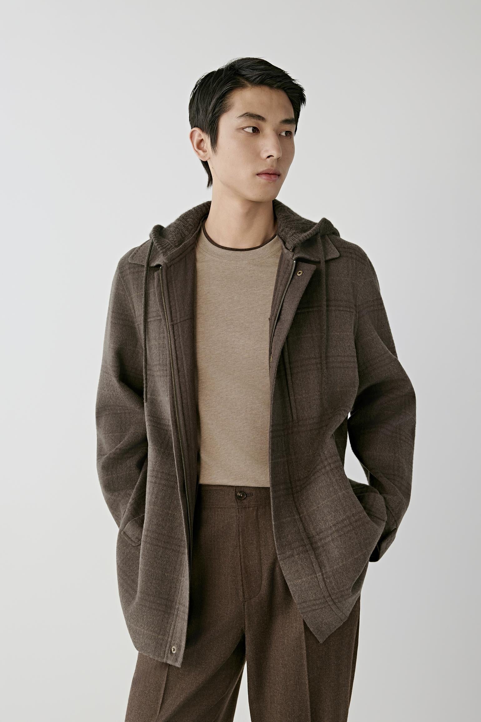 A man wearing a brown plaid hooded jacket over a beige knit sweater and matching brown trousers, photographed against a plain light gray studio background.