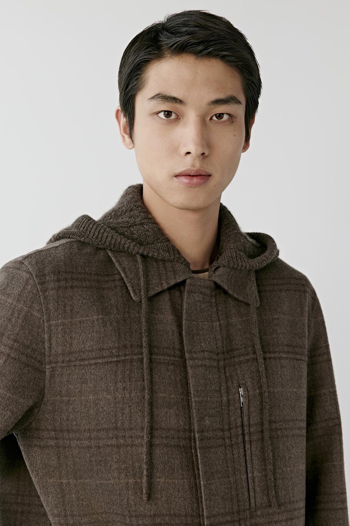 A man wearing a brown textured hooded jacket with a subtle plaid pattern, photographed against a light gray studio background.