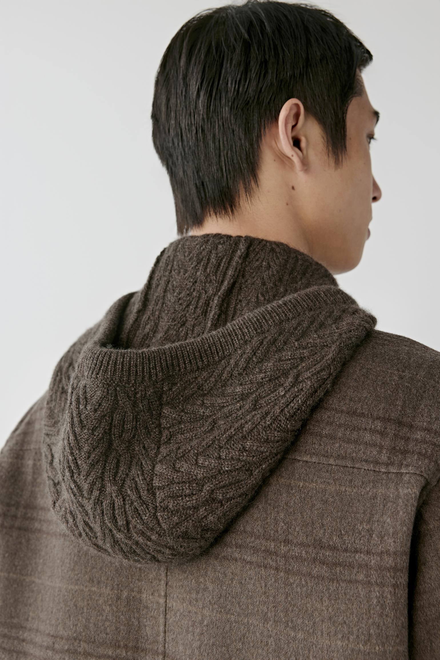 A close-up back view of a person wearing a brown cable-knit hooded sweater layered under a brown plaid coat, against a plain light gray background.