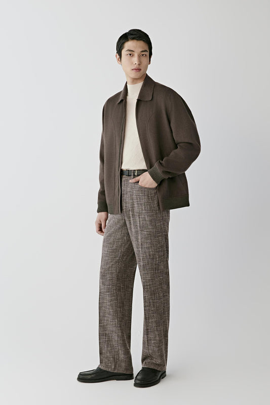 A man modeling a dark brown zip-up jacket over a cream turtleneck, paired with brown and gray plaid trousers and black shoes, against a light gray studio background.