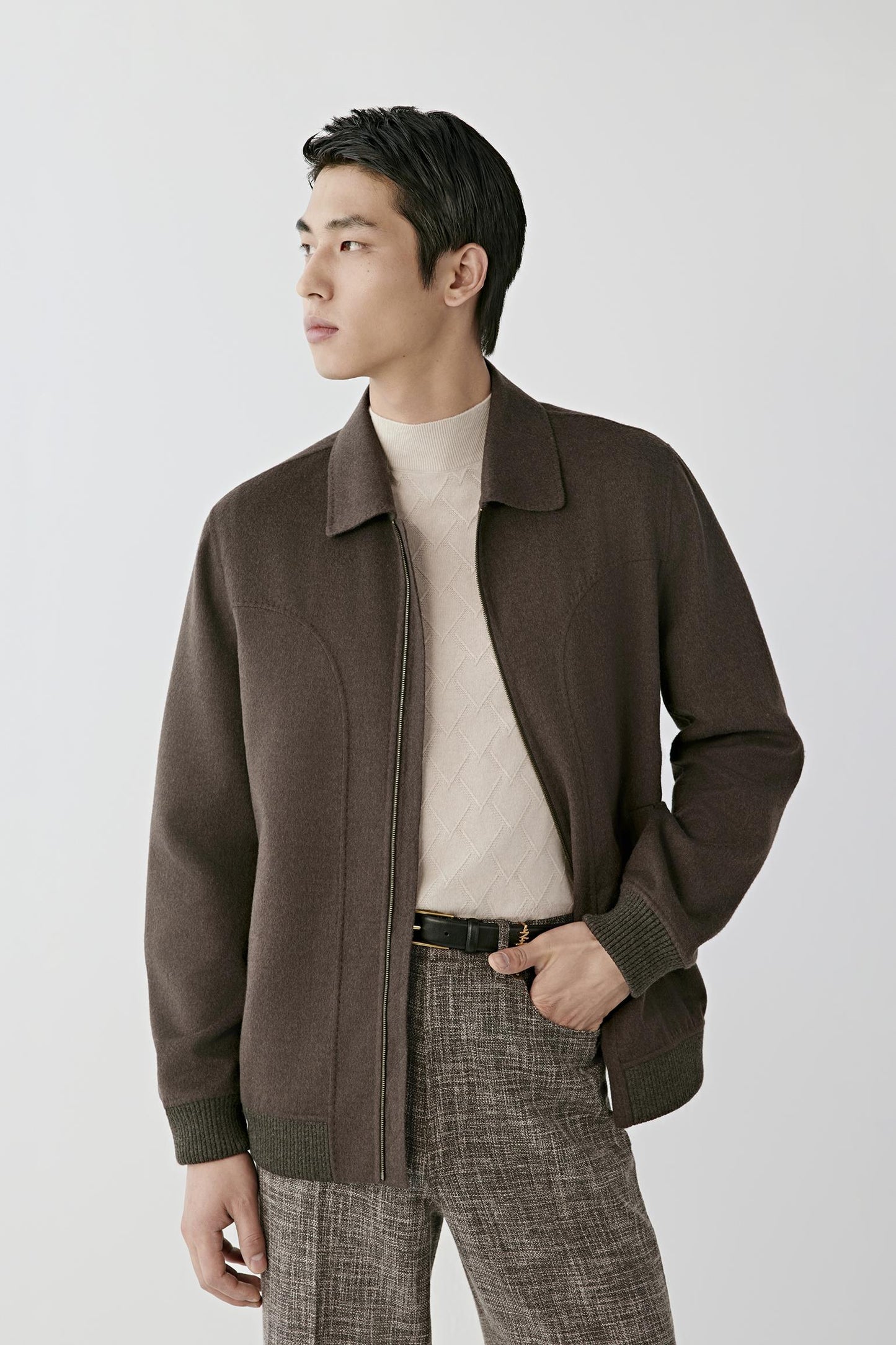 A man wearing a dark brown zip-up jacket over a cream ribbed turtleneck sweater and gray plaid trousers, standing against a light gray studio background.