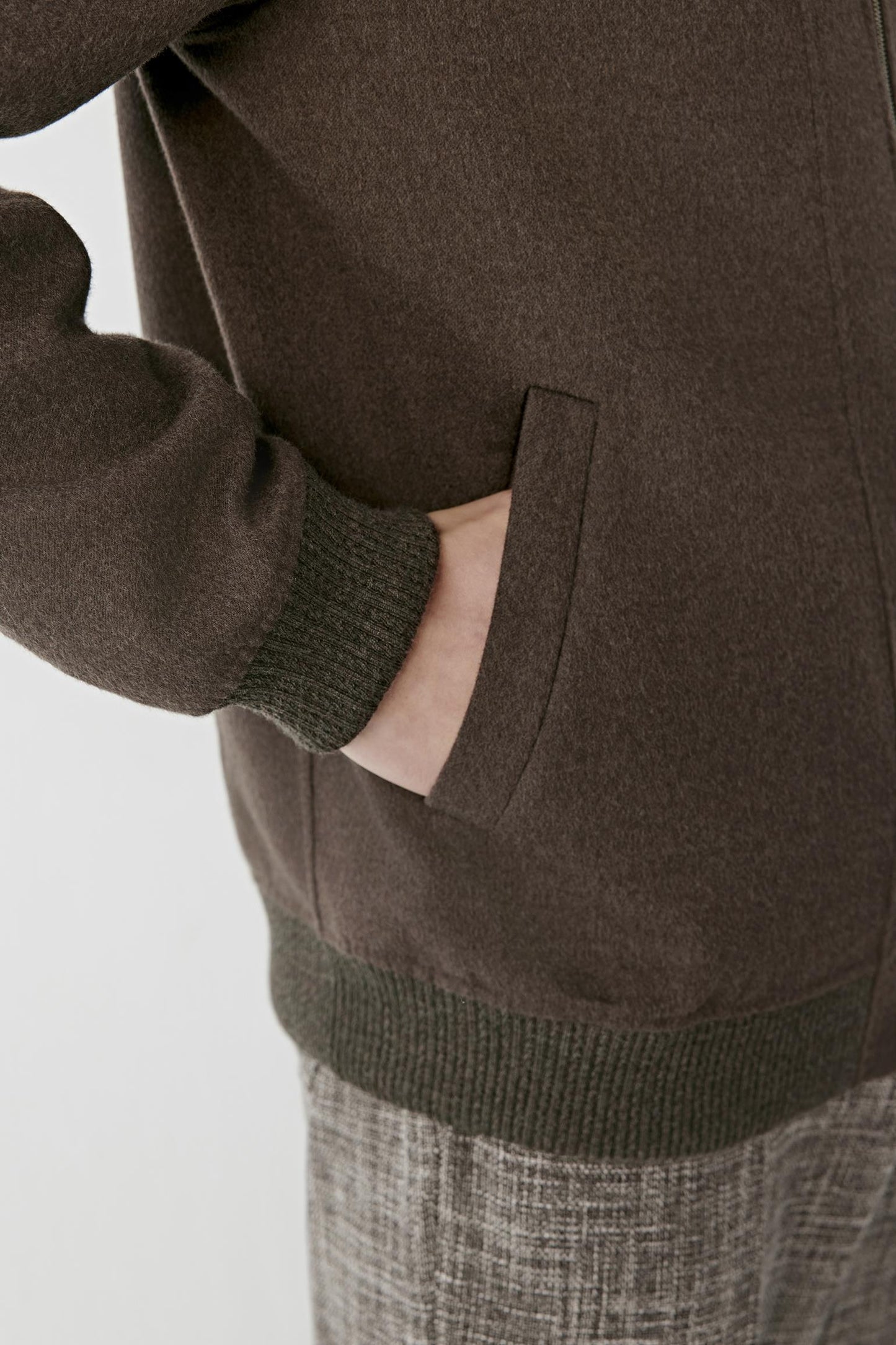 Close-up of a person wearing a dark brown knit sweater with ribbed cuffs and hem, showing a hand in a pocket, paired with gray textured trousers against a light gray background.