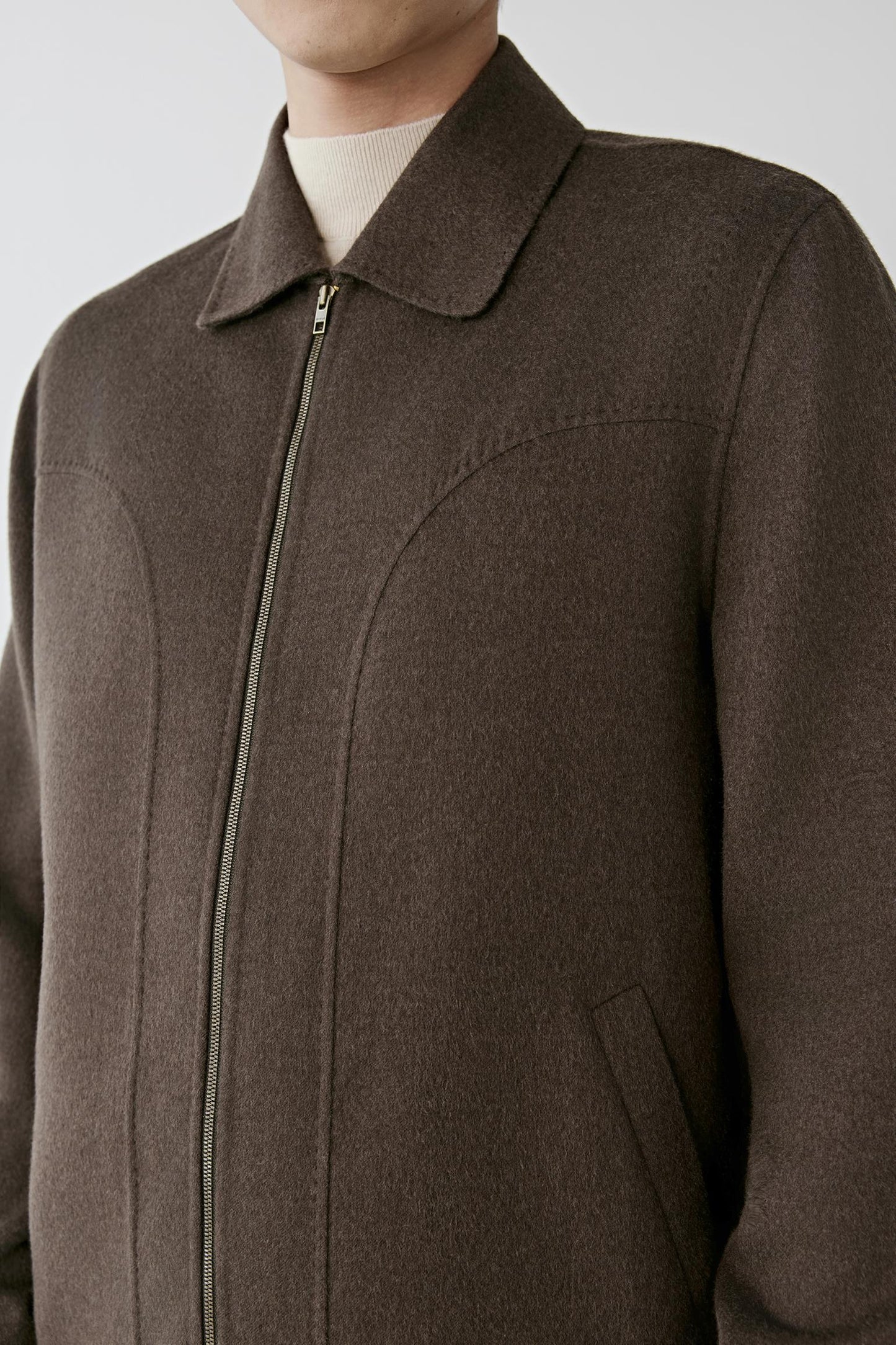 A close-up of a person wearing a dark brown wool-blend jacket with a collar, front zipper, and a light cream t-shirt underneath, set against a plain light gray background.