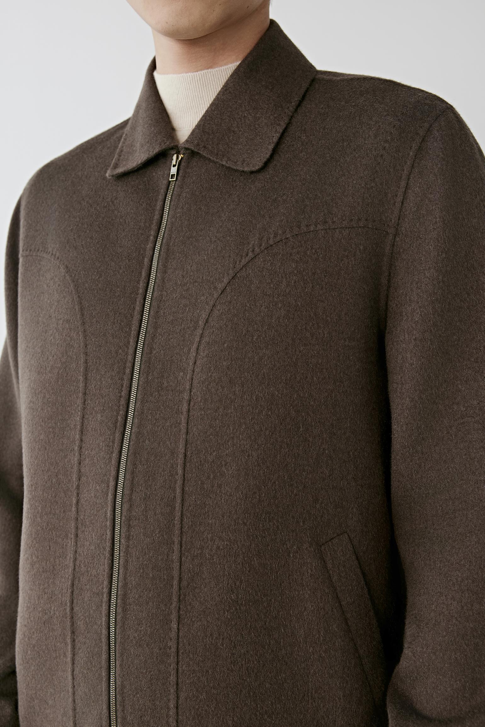 A close-up of a person wearing a dark brown wool-blend jacket with a collar, front zipper, and a light cream t-shirt underneath, set against a plain light gray background.