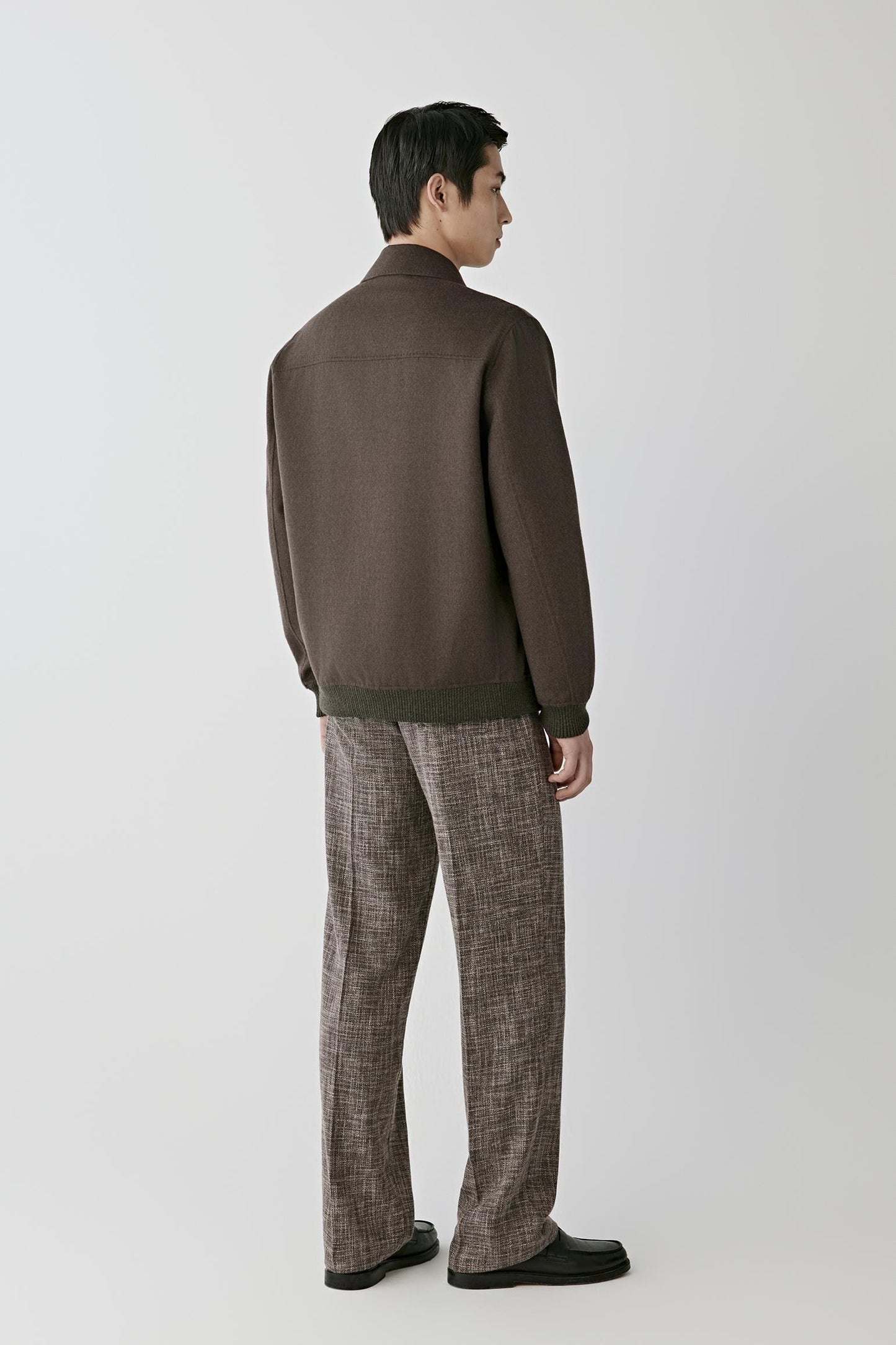 A man shown from the back wearing a dark brown knit jacket with contrasting dark green ribbed trim at the collar, cuffs, and hem, paired with brown and gray patterned trousers and black shoes, against a light gray studio background.