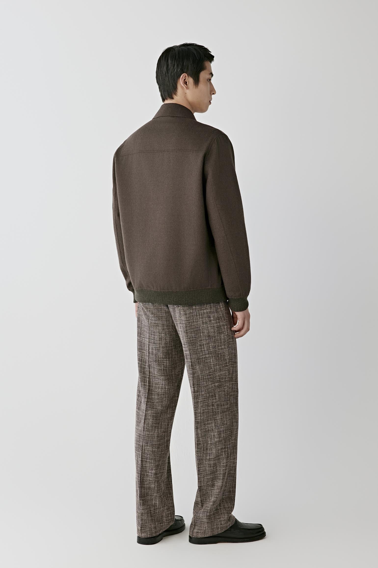 A man shown from the back wearing a dark brown knit jacket with contrasting dark green ribbed trim at the collar, cuffs, and hem, paired with brown and gray patterned trousers and black shoes, against a light gray studio background.