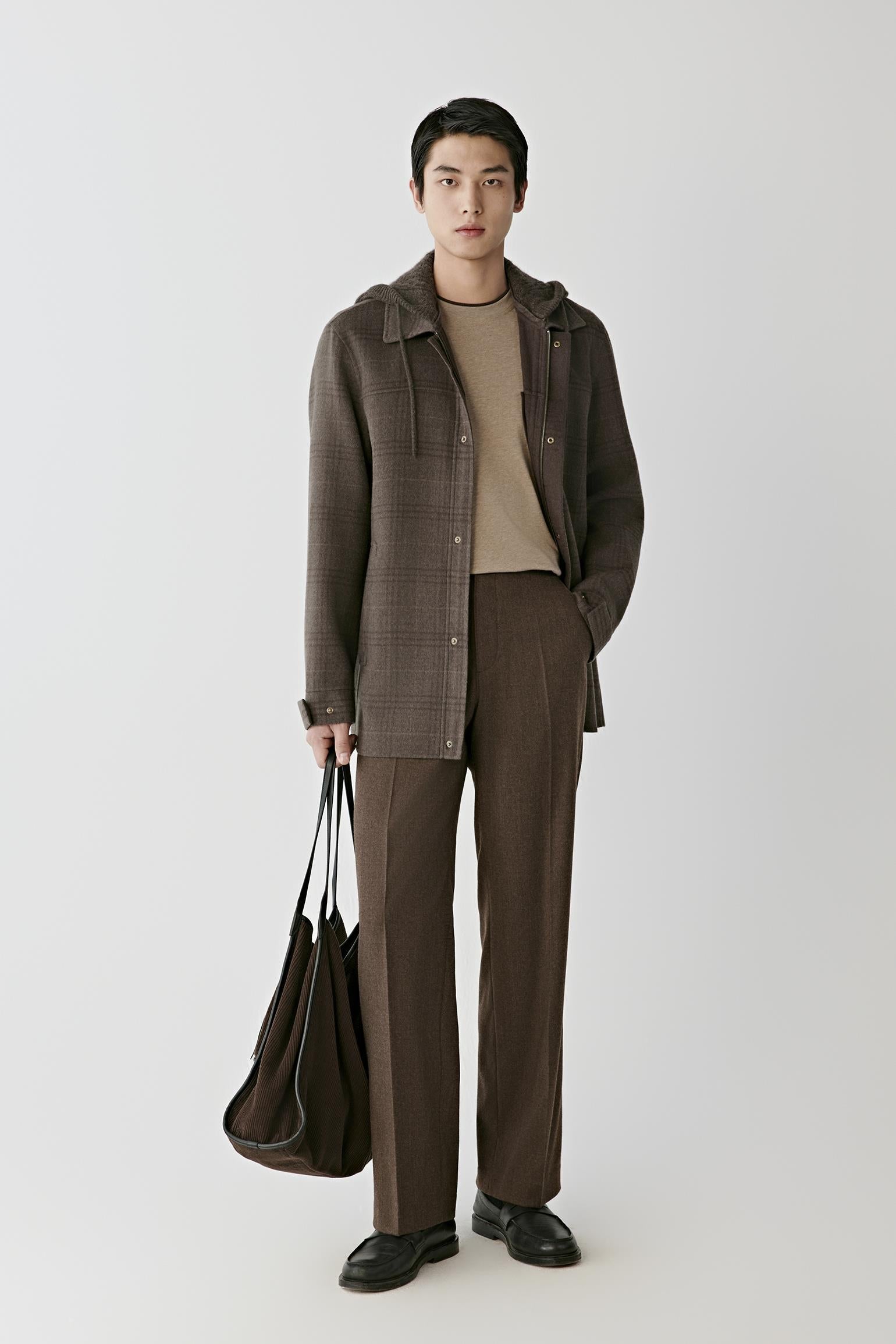 A model stands against a light gray studio background wearing a brown textured hooded jacket over a beige turtleneck, matching brown trousers, and black shoes, while holding a dark brown tote bag.