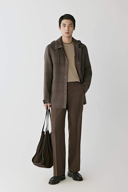 A model stands against a light gray studio background wearing a brown textured hooded jacket over a beige turtleneck, matching brown trousers, and black shoes, while holding a dark brown tote bag.