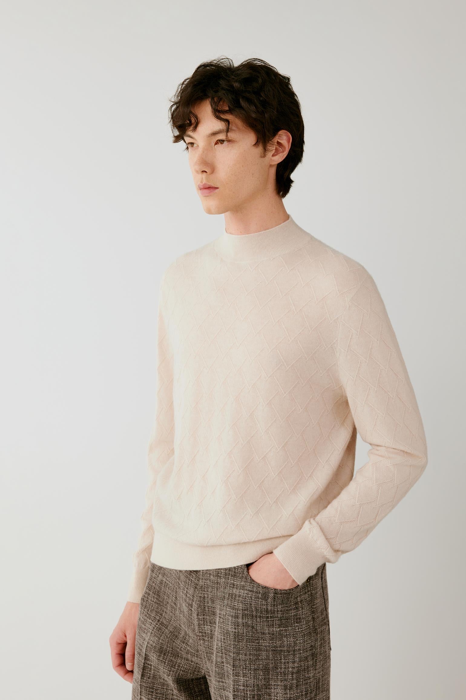 A man wearing a light beige knit mock-neck sweater and textured brown trousers, standing against a plain light gray studio background.