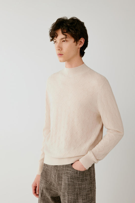 A man wearing a light beige knit mock-neck sweater and textured brown trousers, standing against a plain light gray studio background.