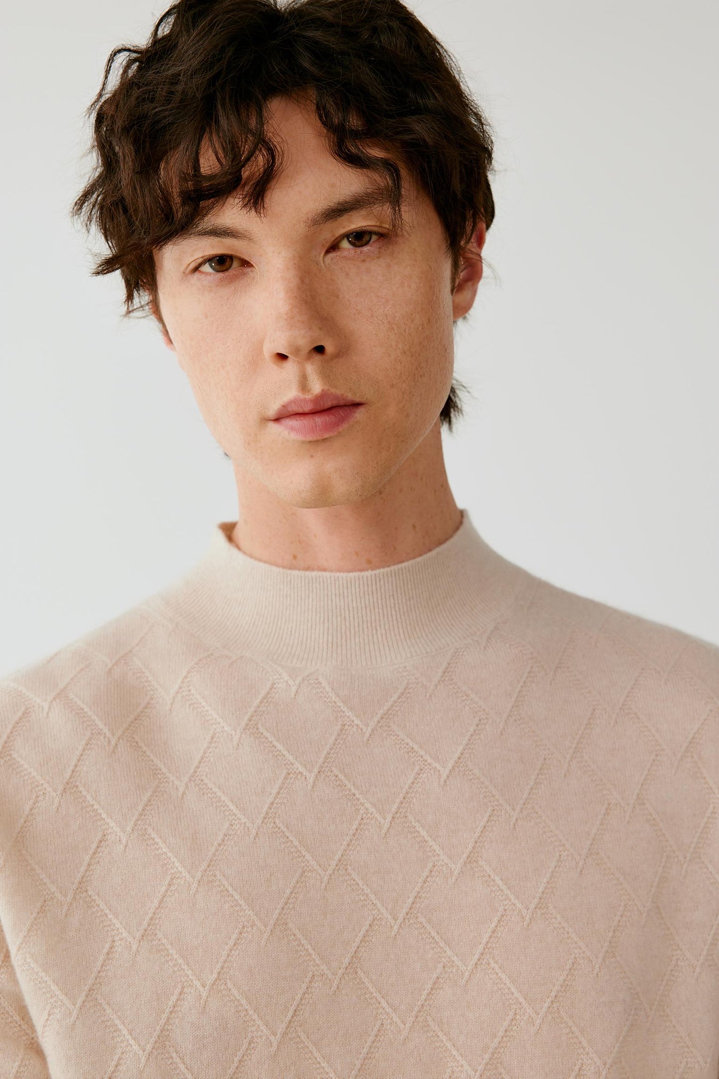A close-up portrait of a man wearing a light beige textured knit mock-neck sweater against a plain light gray background.