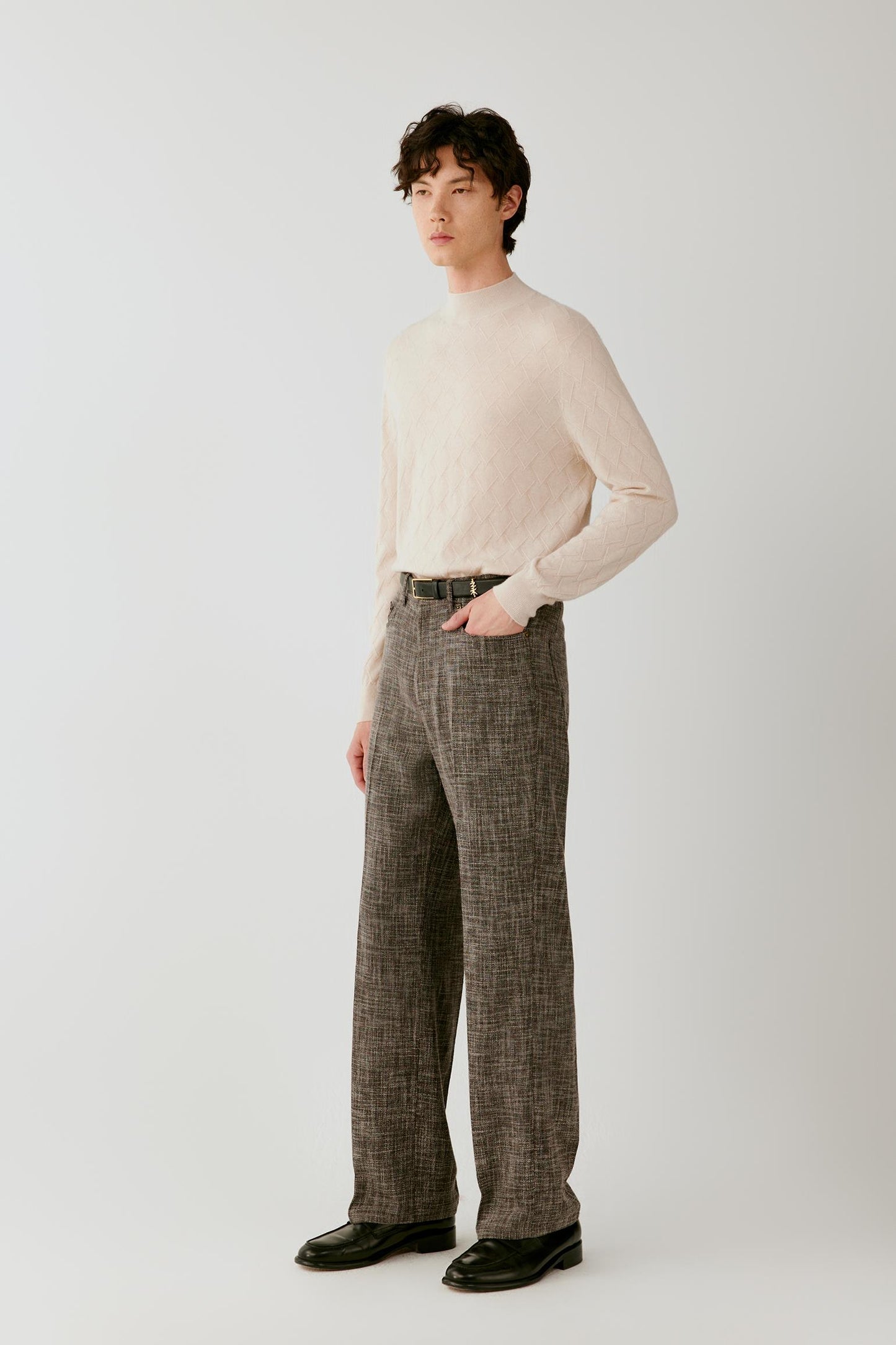 A man modeling a cream ribbed mock-neck sweater and brown plaid wide-leg trousers with black leather shoes, standing against a light gray studio background.