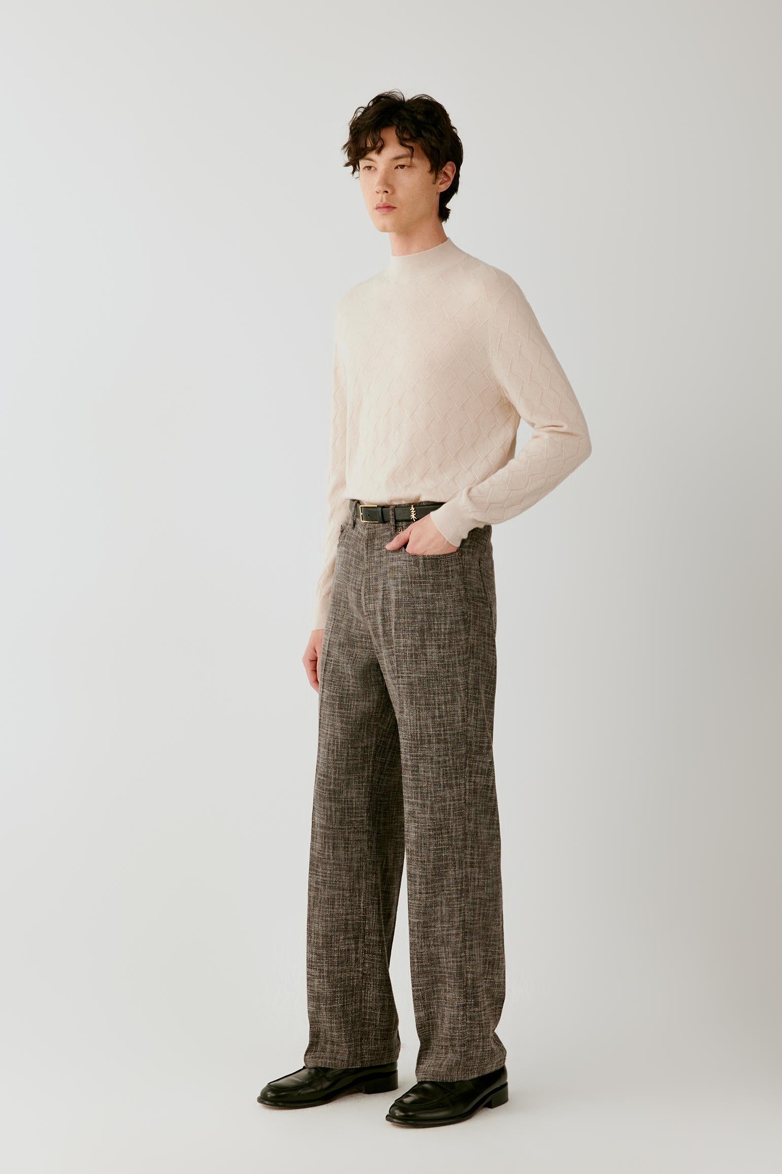 A man modeling a cream ribbed mock-neck sweater and brown plaid wide-leg trousers with black leather shoes, standing against a light gray studio background.