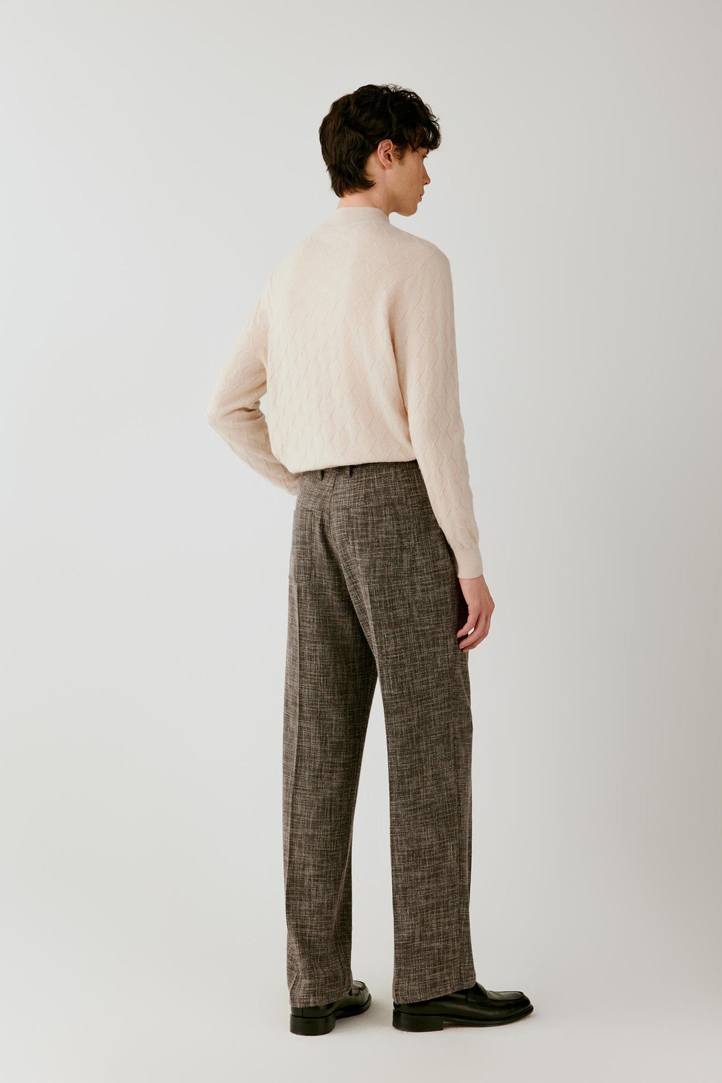 A man shown from the back wearing a cream ribbed knit mock-neck sweater and brown plaid trousers, standing against a light gray studio background.