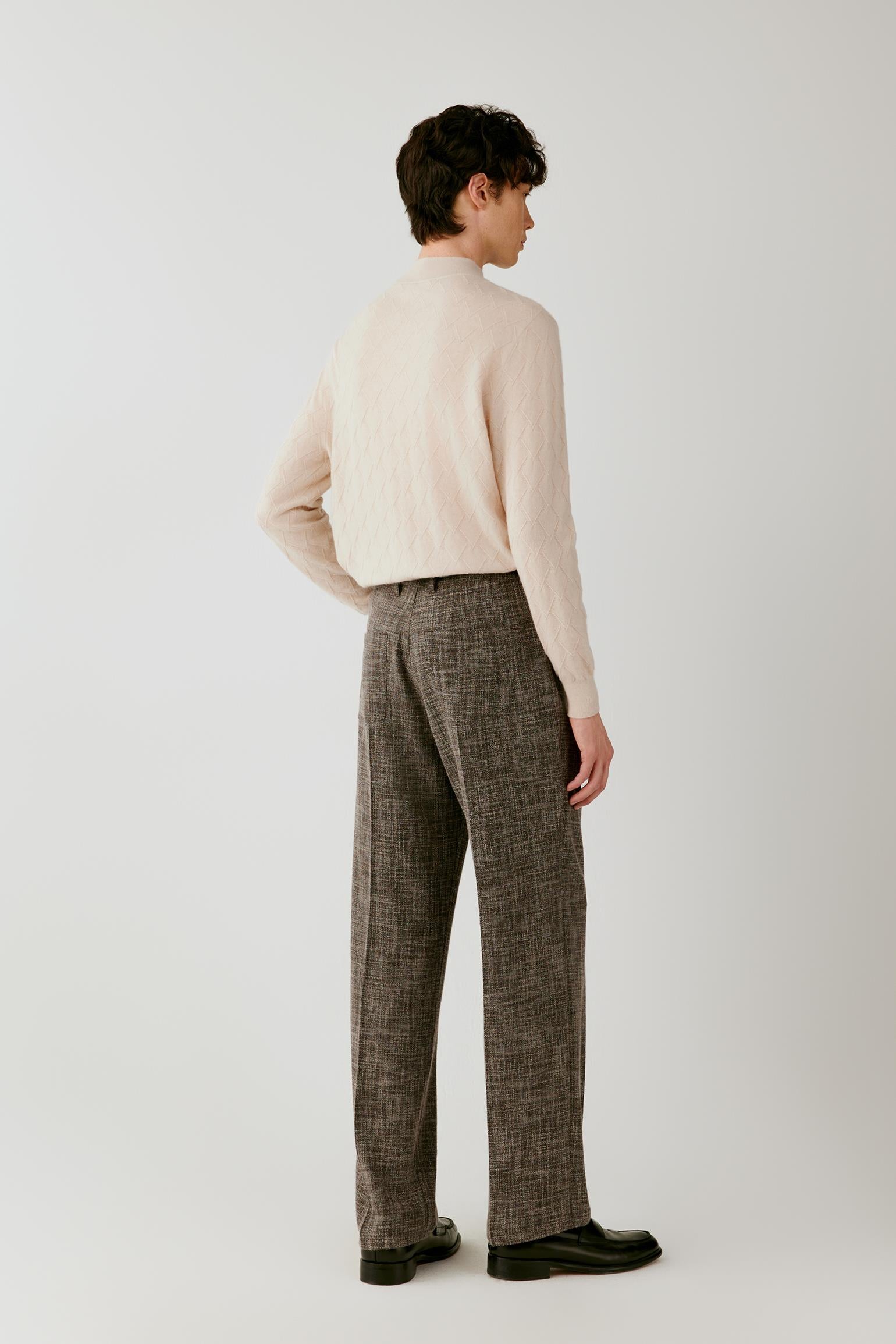 A man shown from the back wearing a cream ribbed knit mock-neck sweater and brown plaid trousers, standing against a light gray studio background.