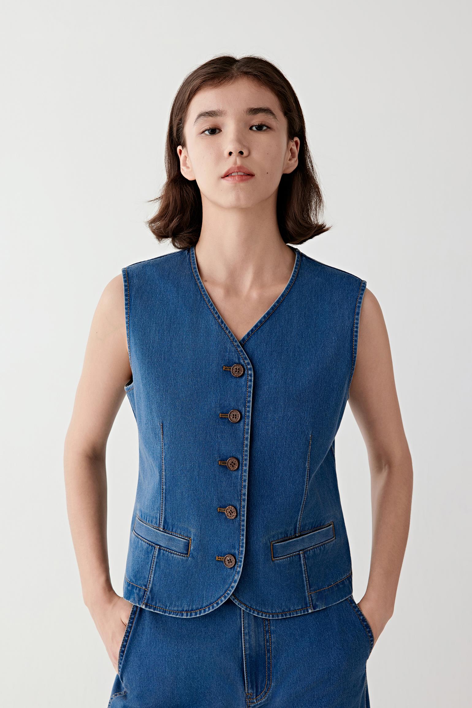 Woman wearing a blue cotton stretch denim vest with a fitted, sleeveless cut and V-neckline, front view showing the buttoned front, defined waist shaping and structured yet flexible denim.
