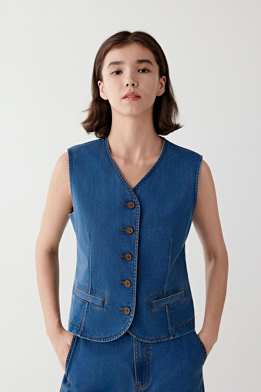 Woman wearing a blue cotton stretch denim vest with a fitted, sleeveless cut and V-neckline, front view showing the buttoned front, defined waist shaping and structured yet flexible denim.