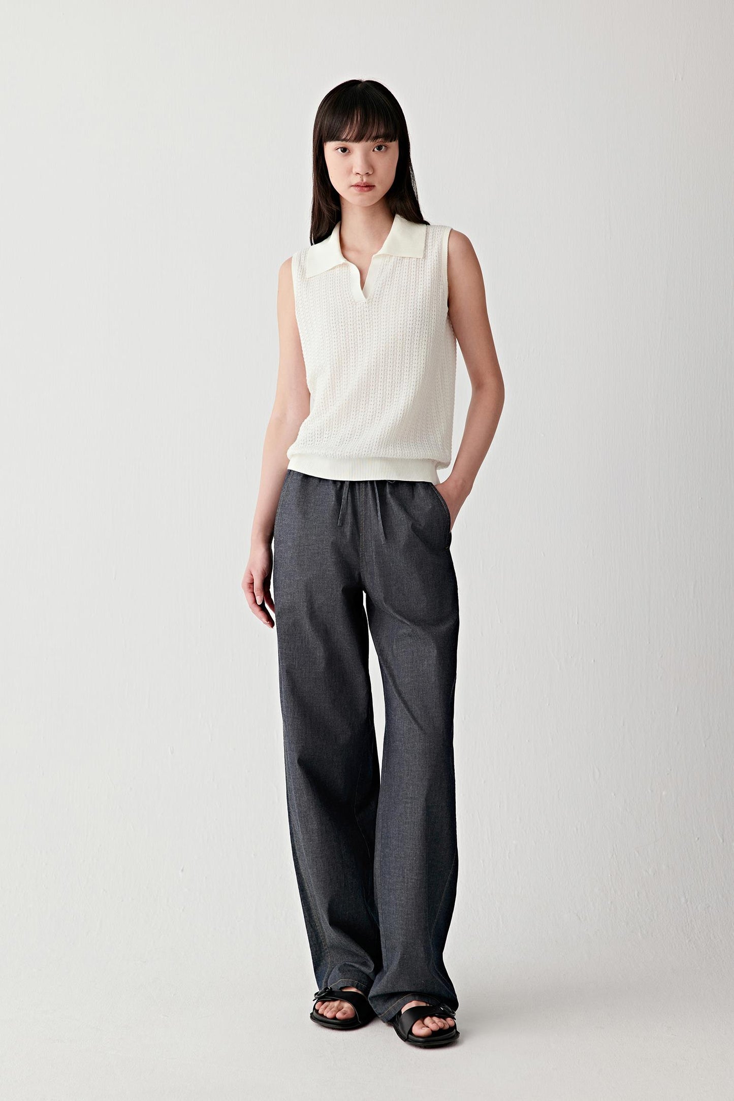 Full-length front view of a woman standing facing forward wearing an ivory sleeveless cotton polo, paired with dark wide-leg trousers, highlighting the clean silhouette, soft structure of the knit and the contrast between the light top and darker bottom