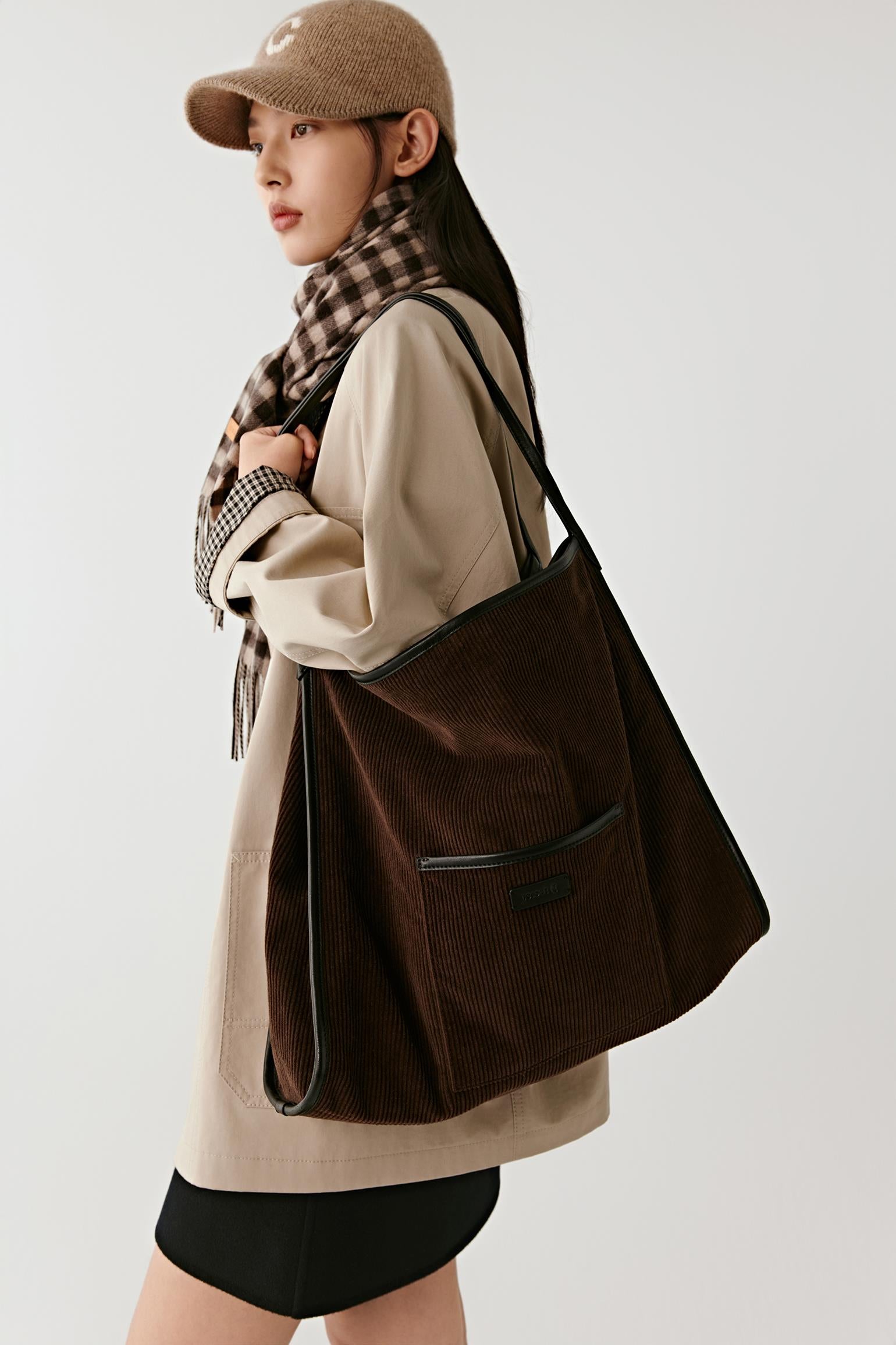 A woman wearing a beige coat, black skirt, brown plaid scarf, and brown cap carries a large dark brown corduroy tote bag with black trim against a light gray studio background.