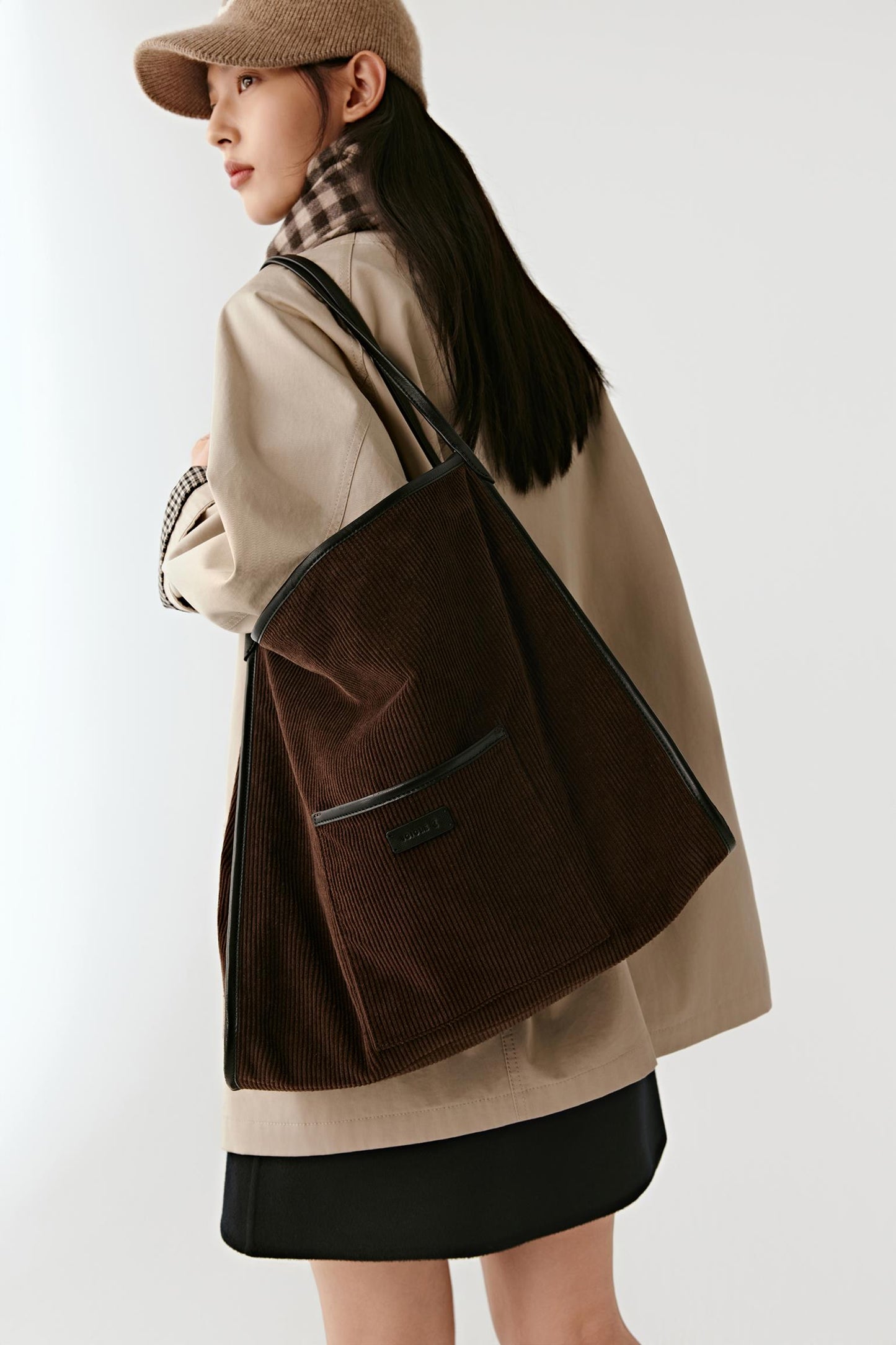A woman wearing a beige coat, black skirt, and brown hat carries a large dark brown corduroy tote bag with black trim over her shoulder, photographed against a plain light gray studio background.